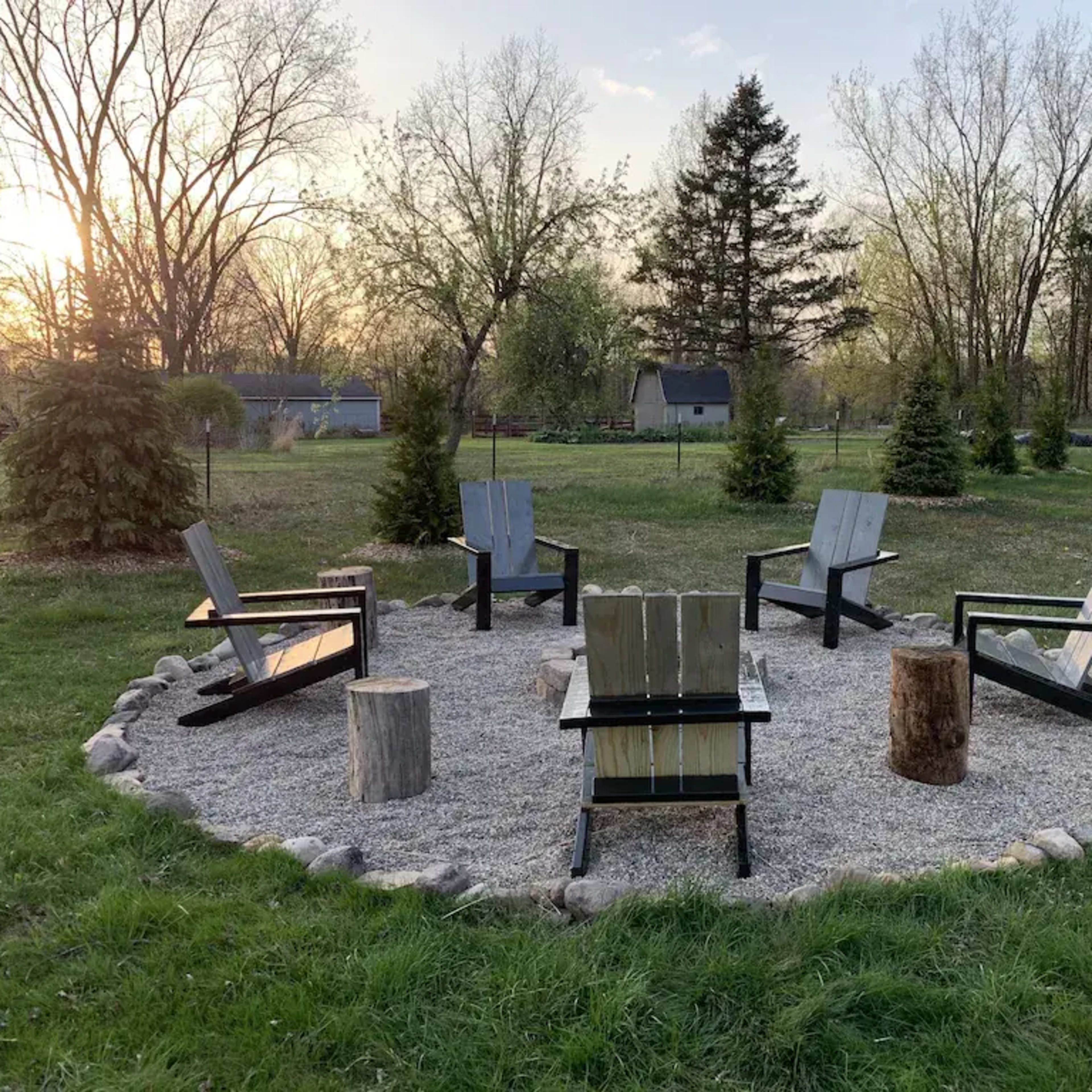 A circular arrangement of six wooden chairs around a gravel area, surrounded by grass and trees, with a sunset in the background.