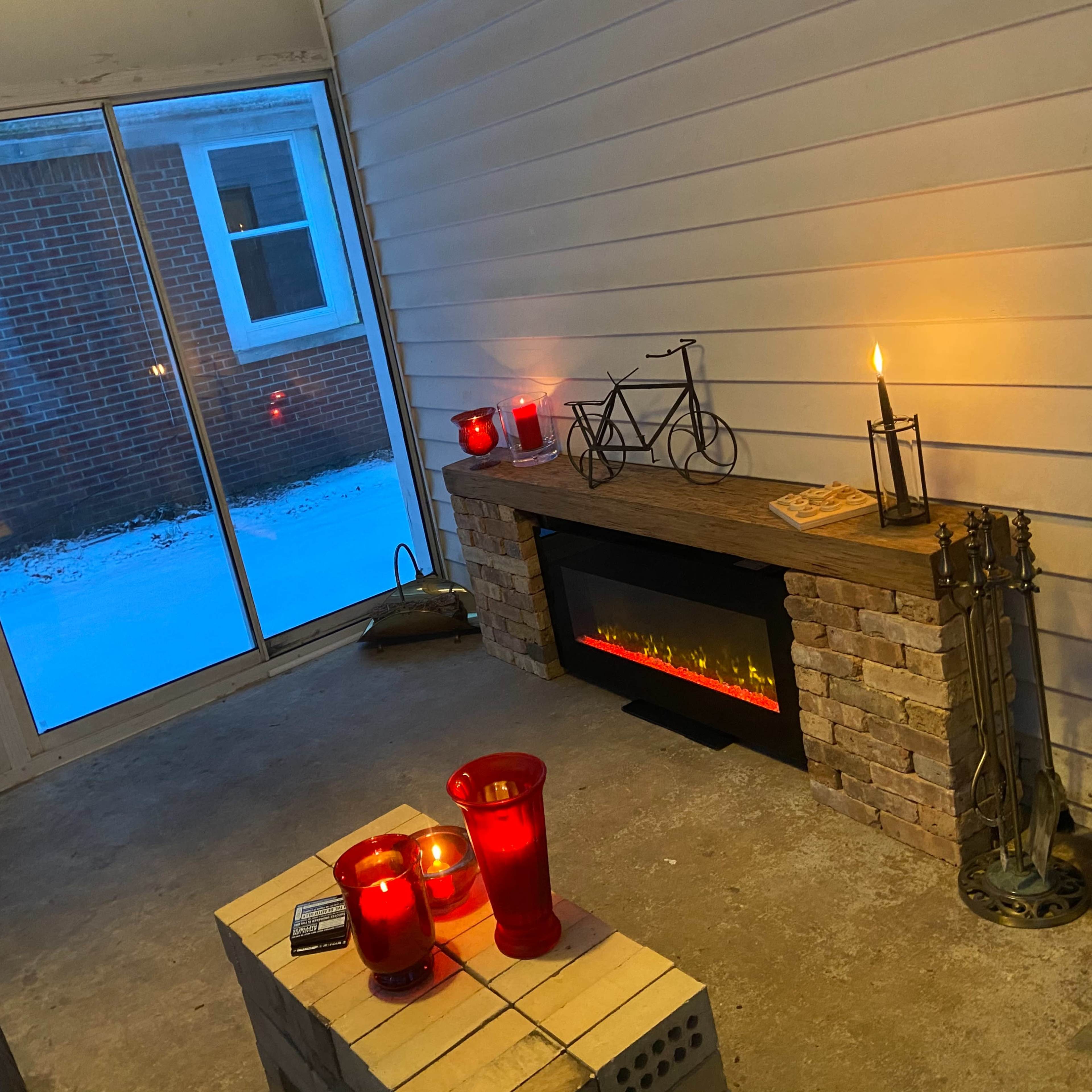 A cozy indoor space with a wooden mantel, candles in red glass holders, an electric fireplace, and a view of snow outside through large glass doors.