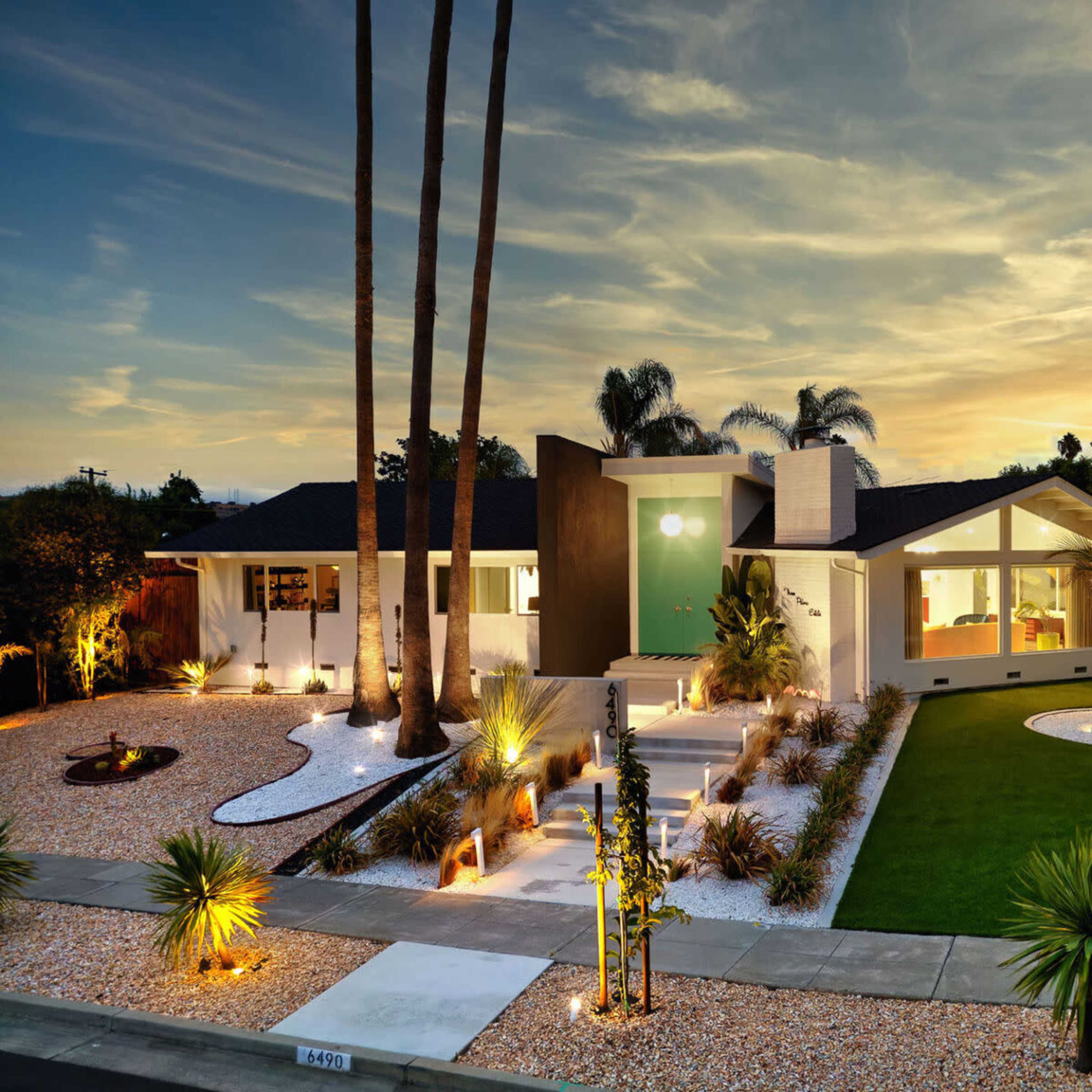 A modern single-story house with large windows and a landscaped yard featuring palm trees and a gravel pathway is illuminated by evening light.