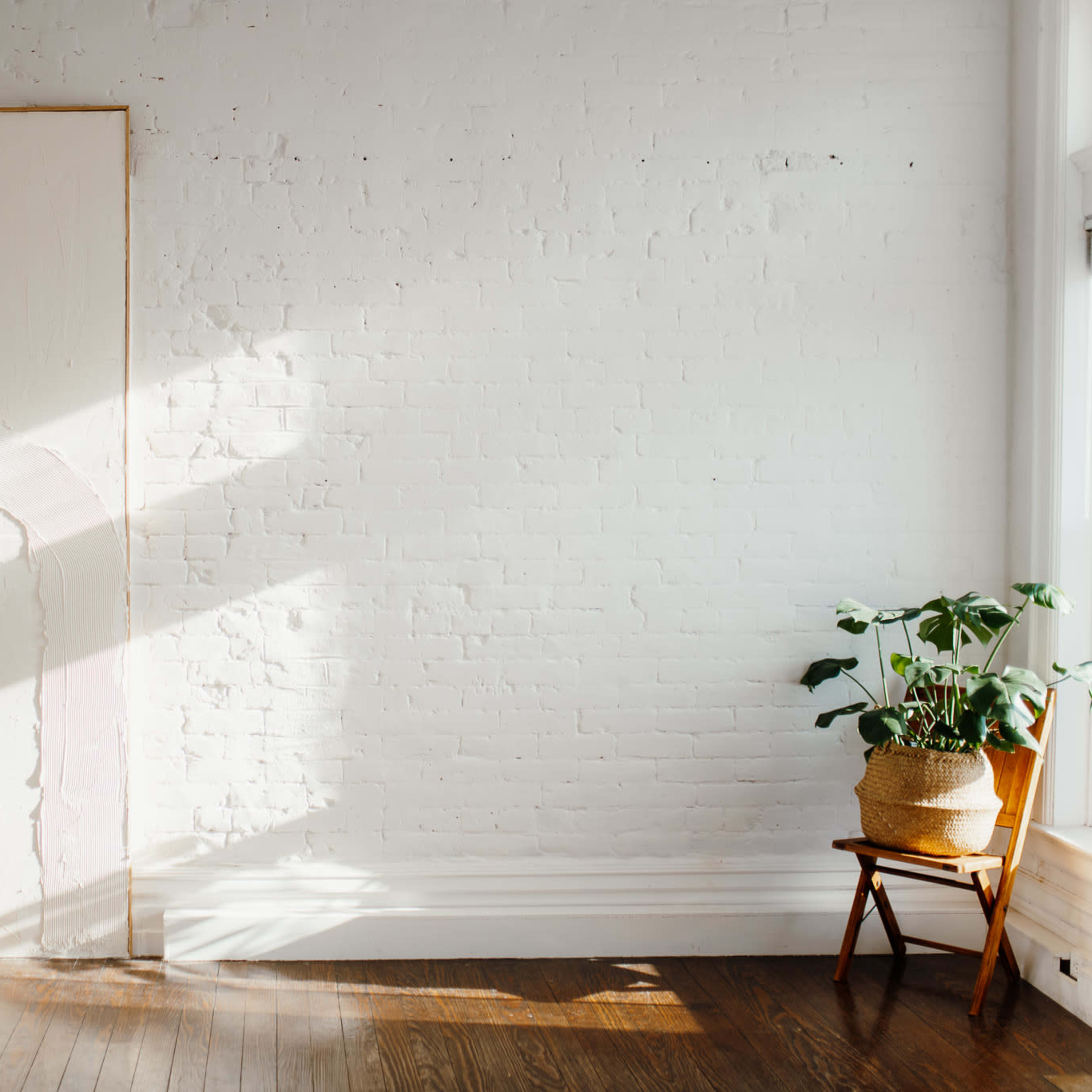 A wooden ladder leans against a white brick wall beside a large, blank canvas, while a potted plant sits next to a wooden chair in a sunlit room.