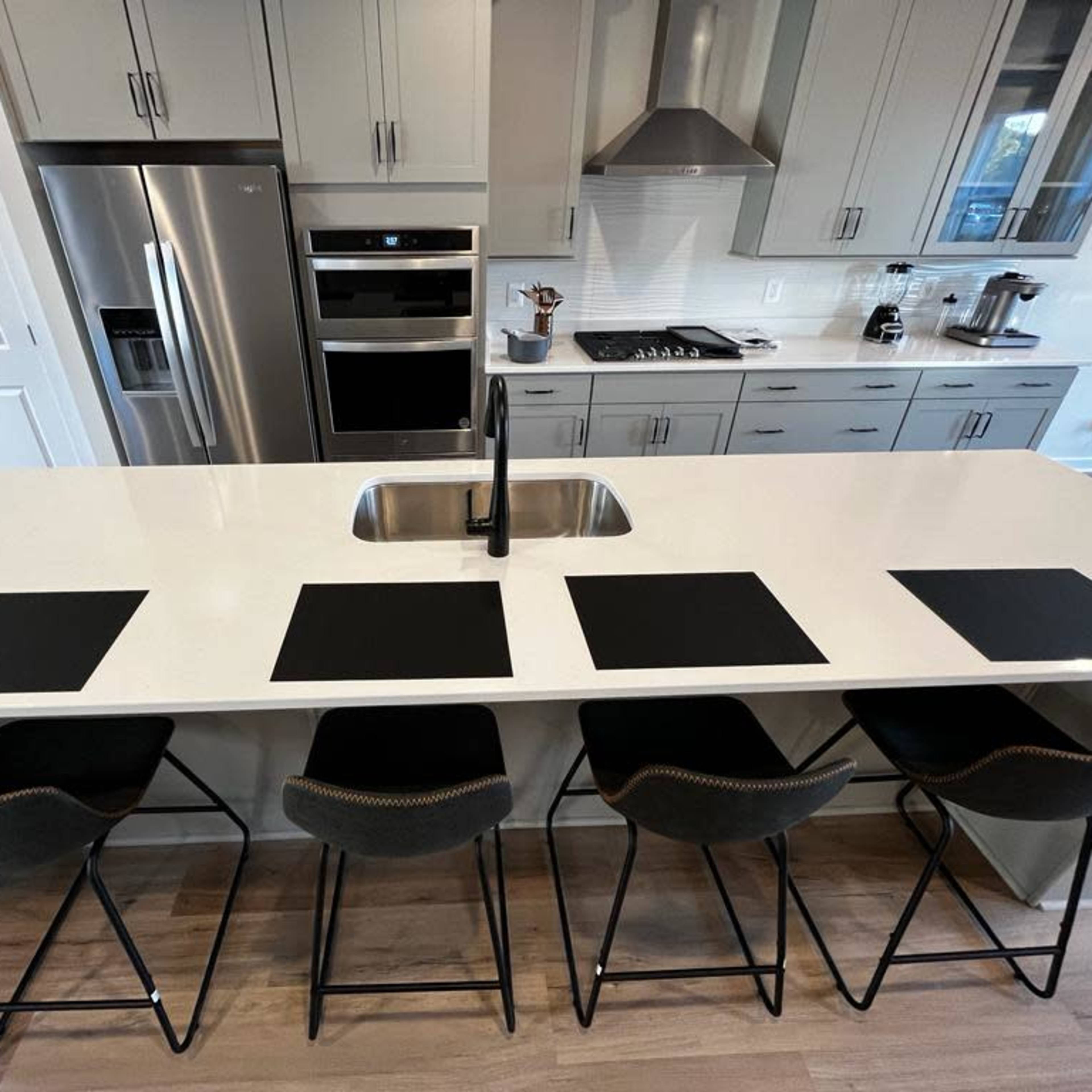 A modern kitchen features a large island with a sink and four black placemats placed in front of stylish bar stools.