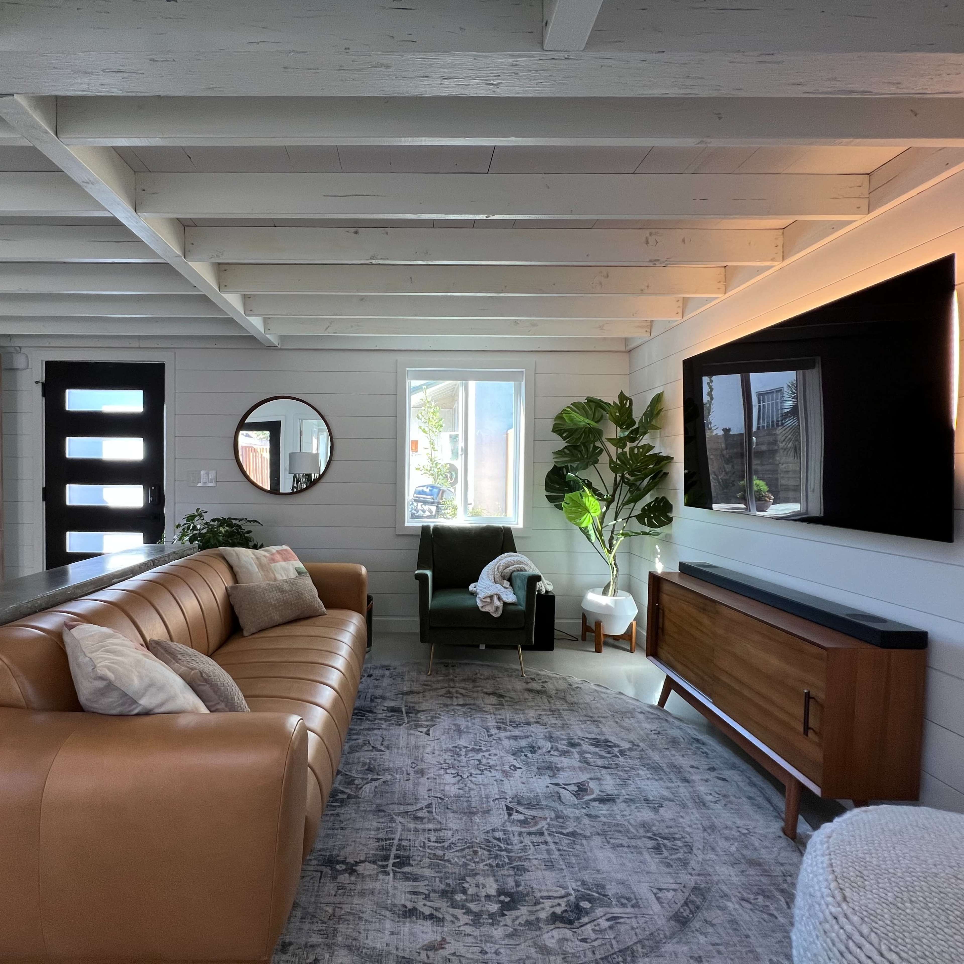 The living room features a tan leather sofa, a dark green armchair, a wooden media console beneath a wall-mounted TV, and a large window beside a round mirror.