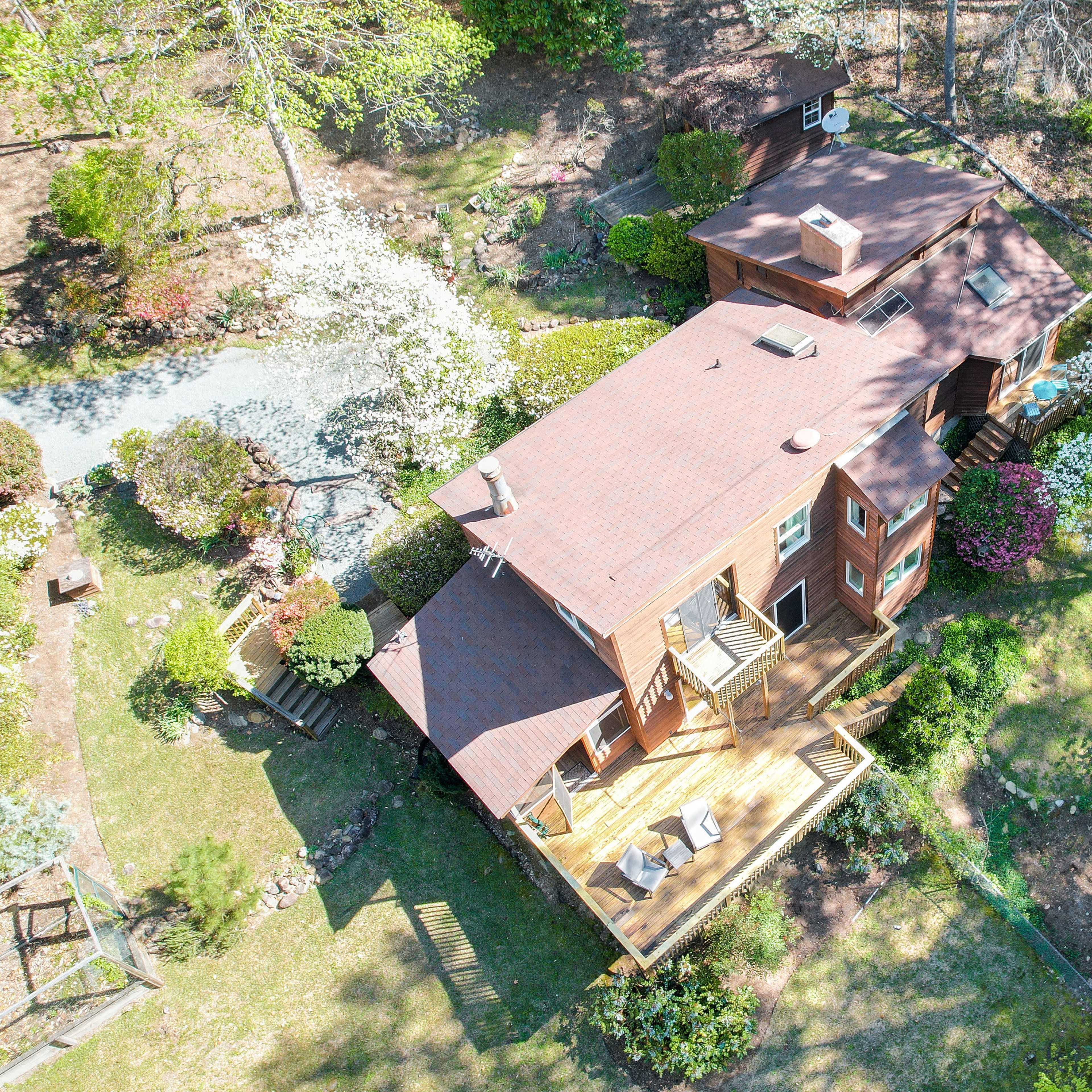 An aerial view shows a wooden house surrounded by trees and flowering plants in a landscaped yard.