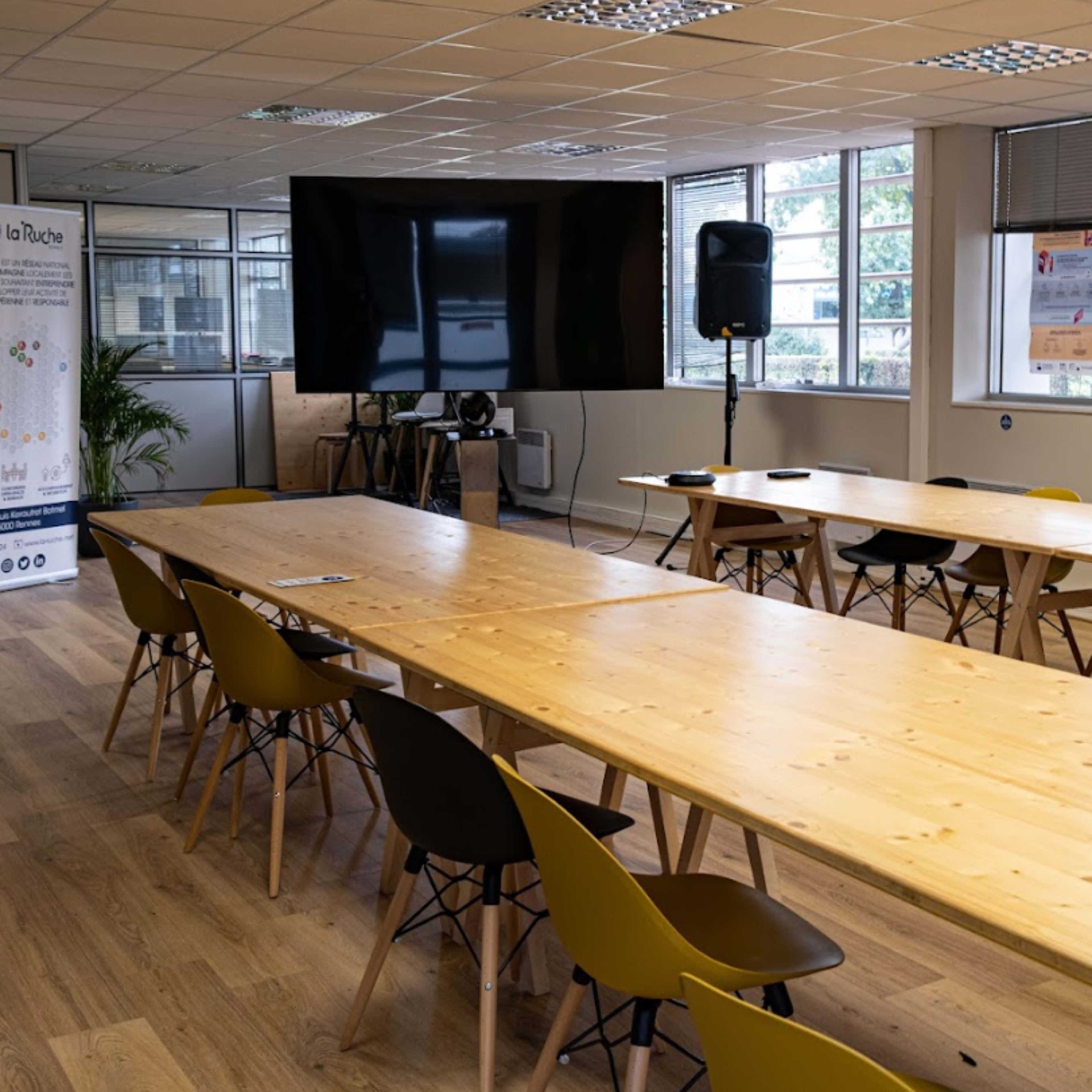 A spacious meeting room features long wooden tables arranged in a U-shape, surrounded by chairs, with large windows allowing natural light and a screen for presentations.