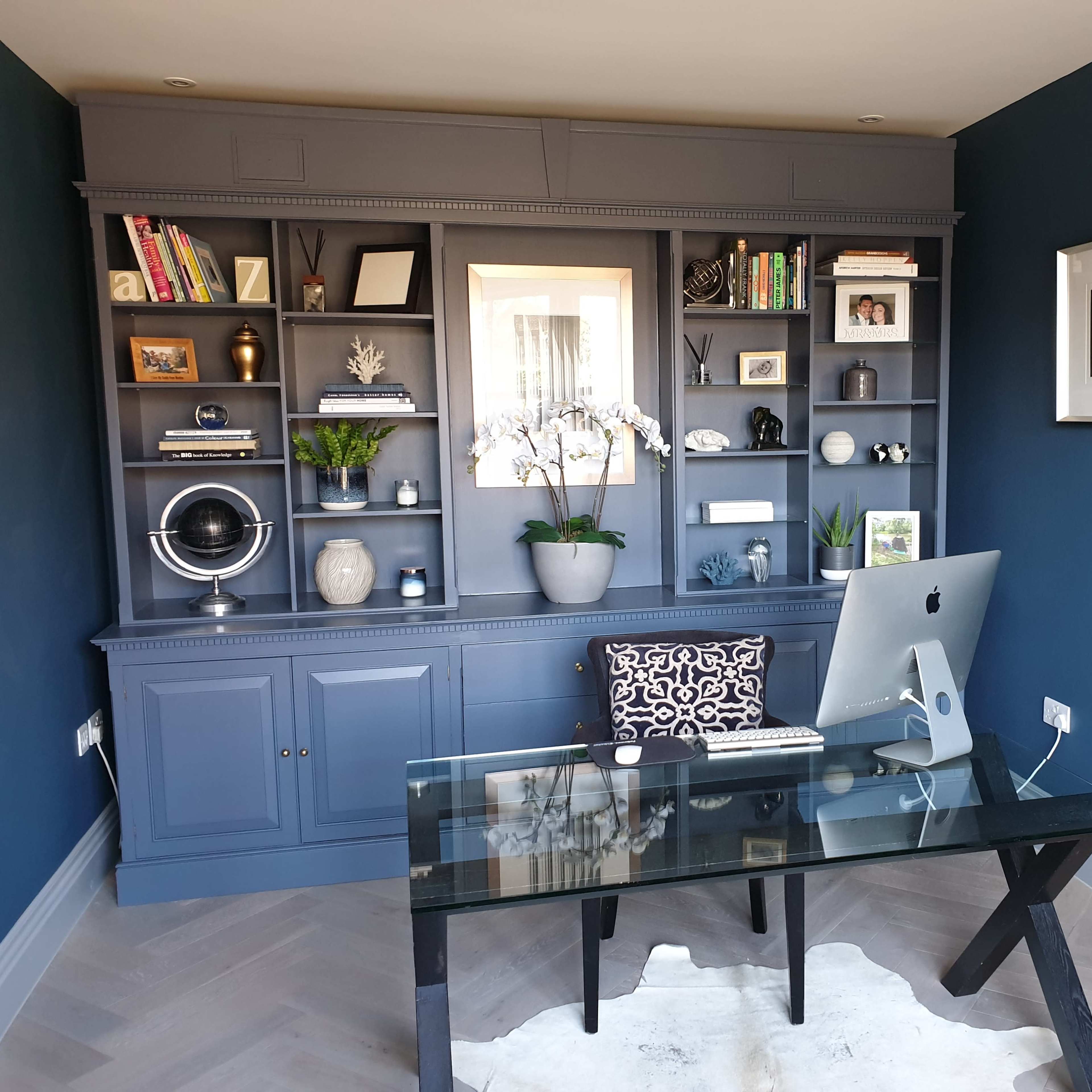 A modern office features a glass desk in front of a dark blue wall with a bookshelf displaying various decor items and plants.