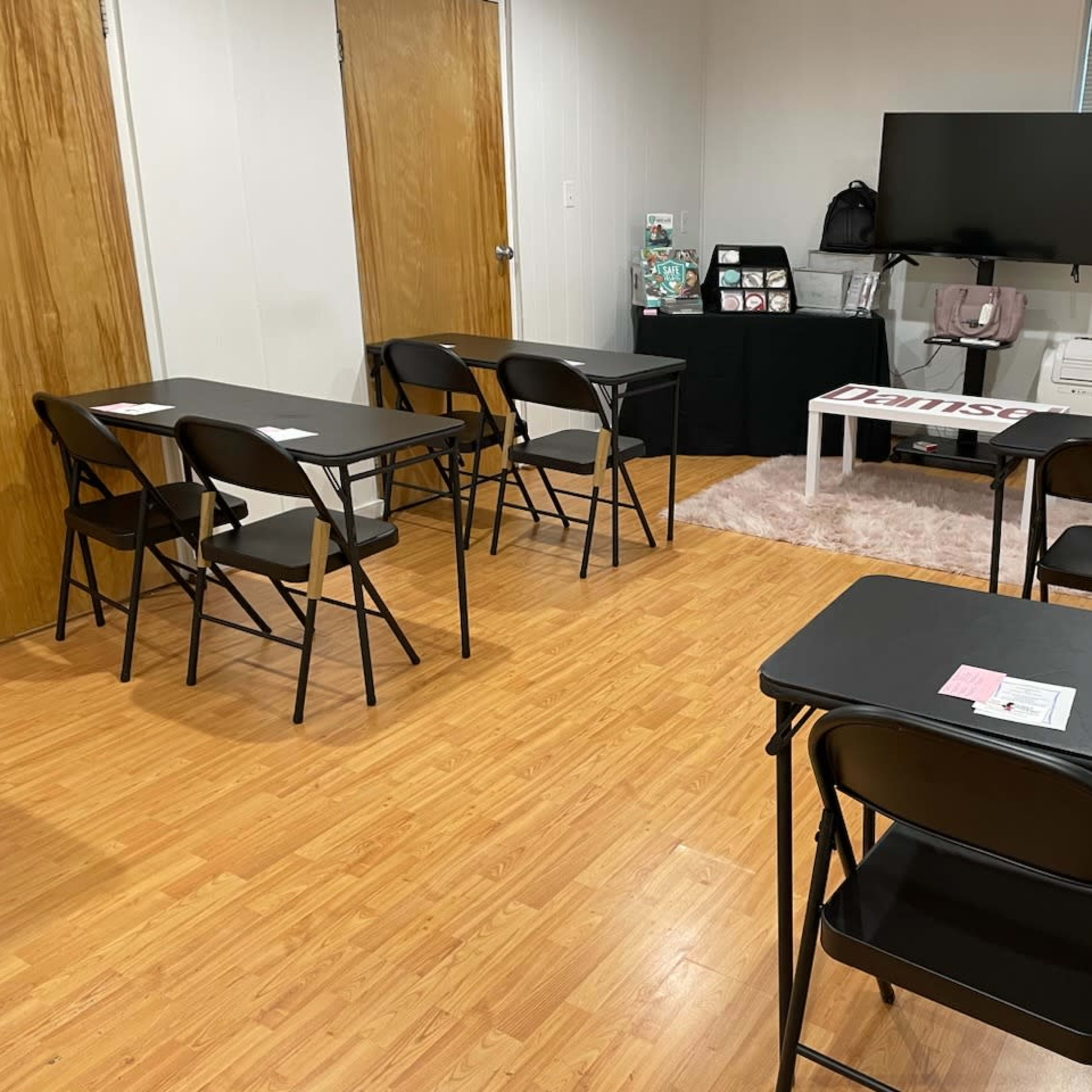 A classroom setup with black folding tables and chairs arranged facing a television in a room with light-colored walls and wooden floors.