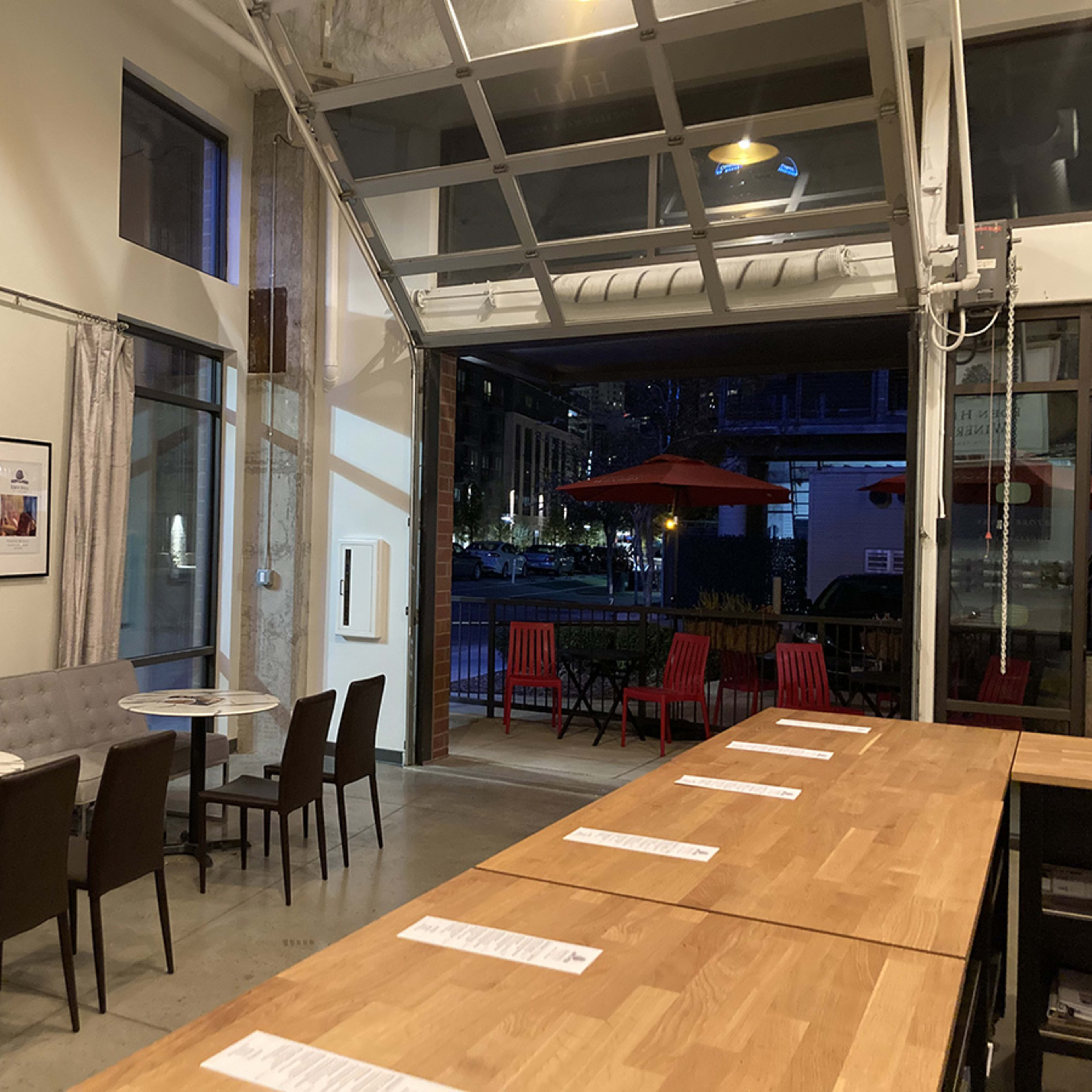 A modern café interior with tables and chairs arranged near an open garage door that leads to an outdoor seating area.