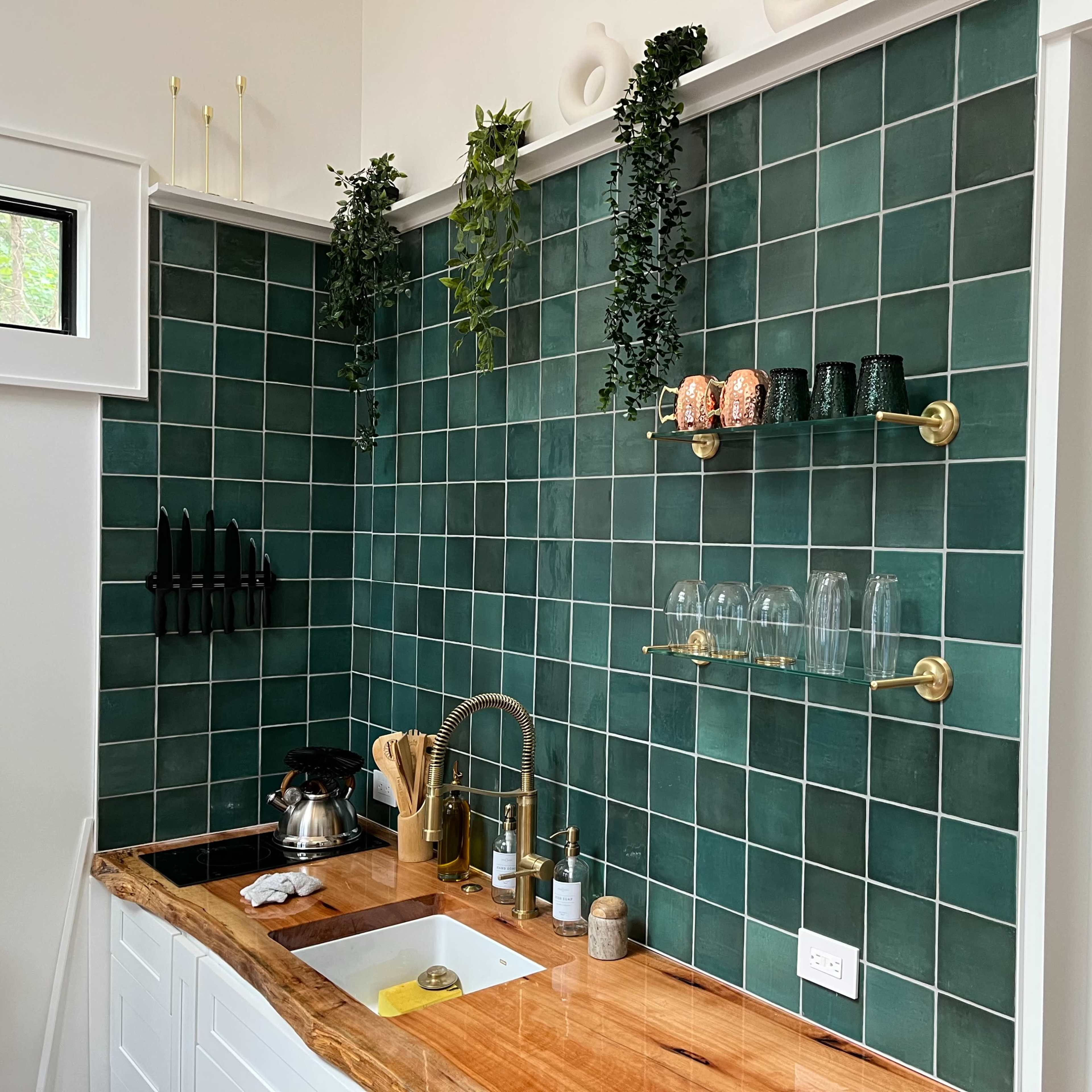 A modern kitchen with green tiled walls, wooden countertops, and shelves displaying glasses and utensils.