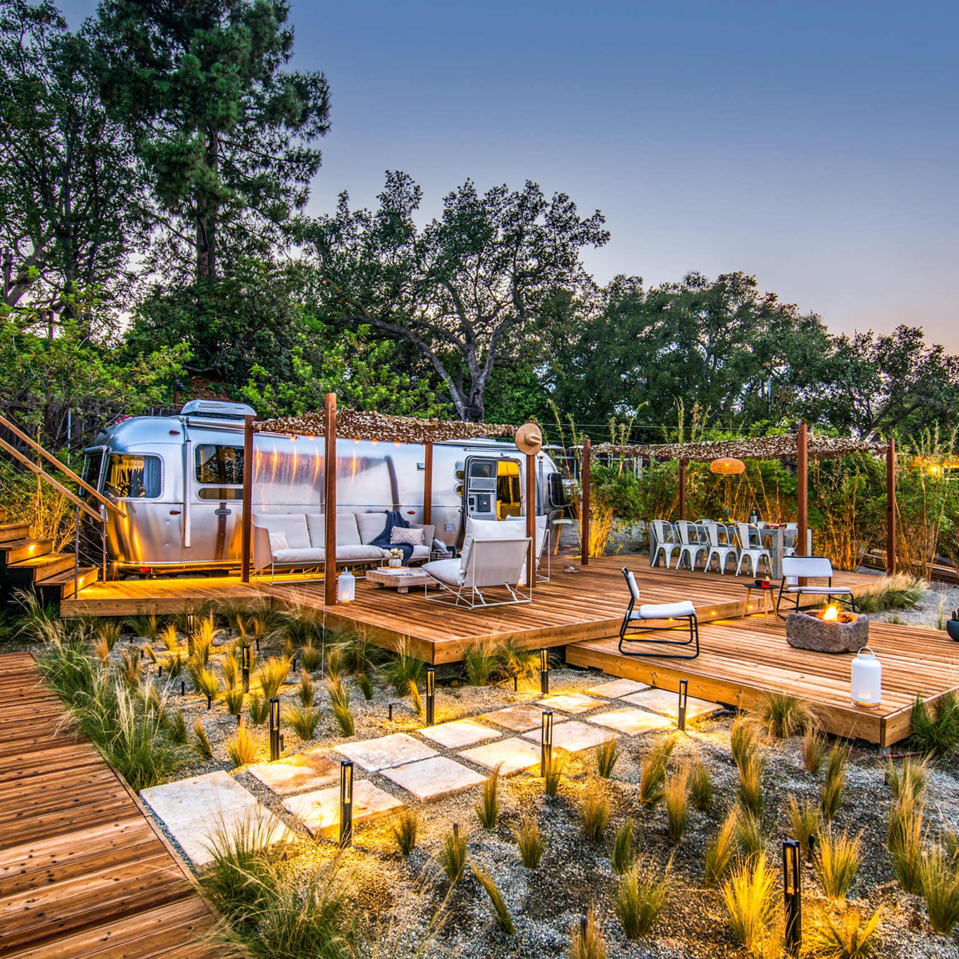 A modern outdoor space features a vintage Airstream trailer, surrounded by wooden decking, seating areas, and landscaped greenery.