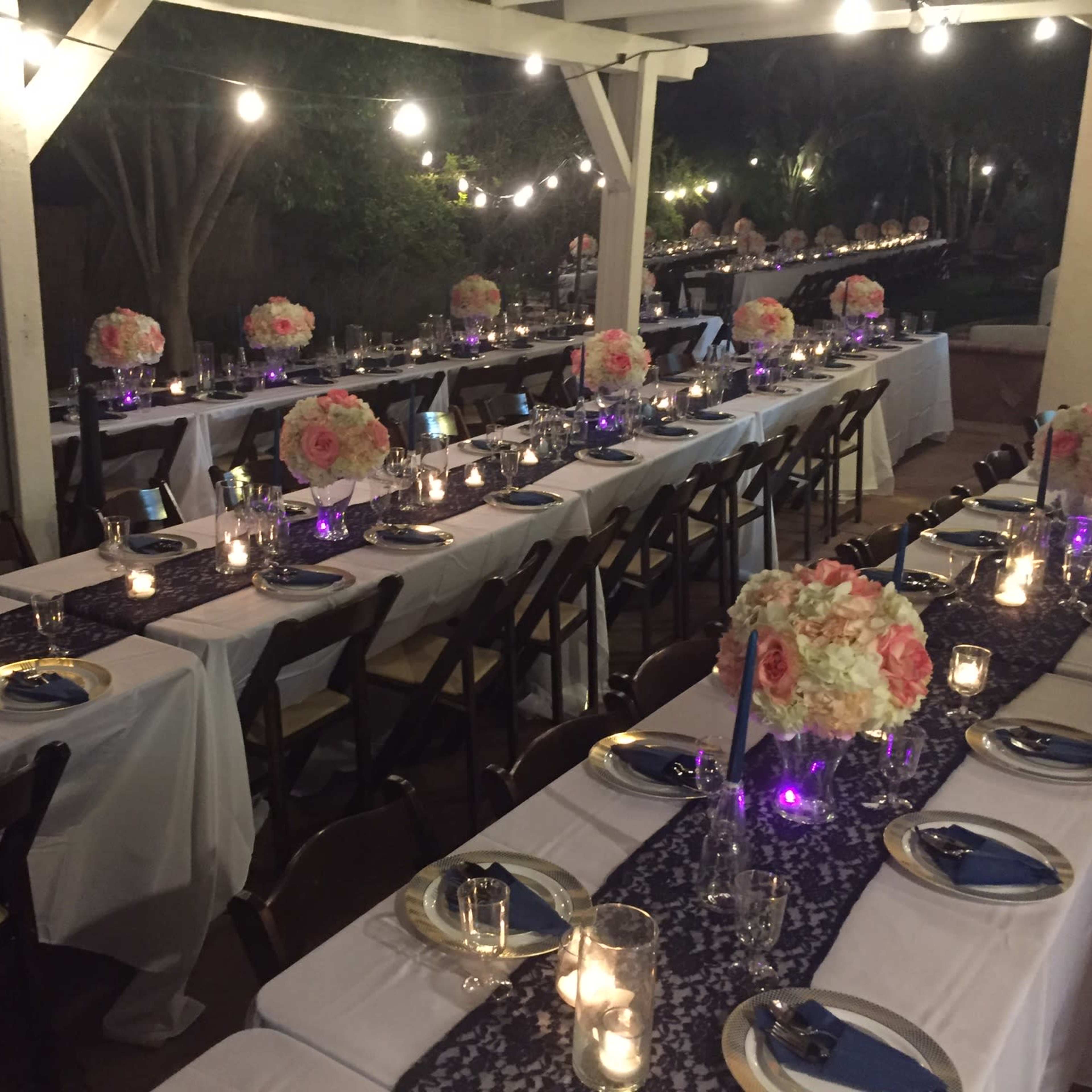 A row of elegantly set tables adorned with floral centerpieces and candlelight is arranged under string lights on a patio.