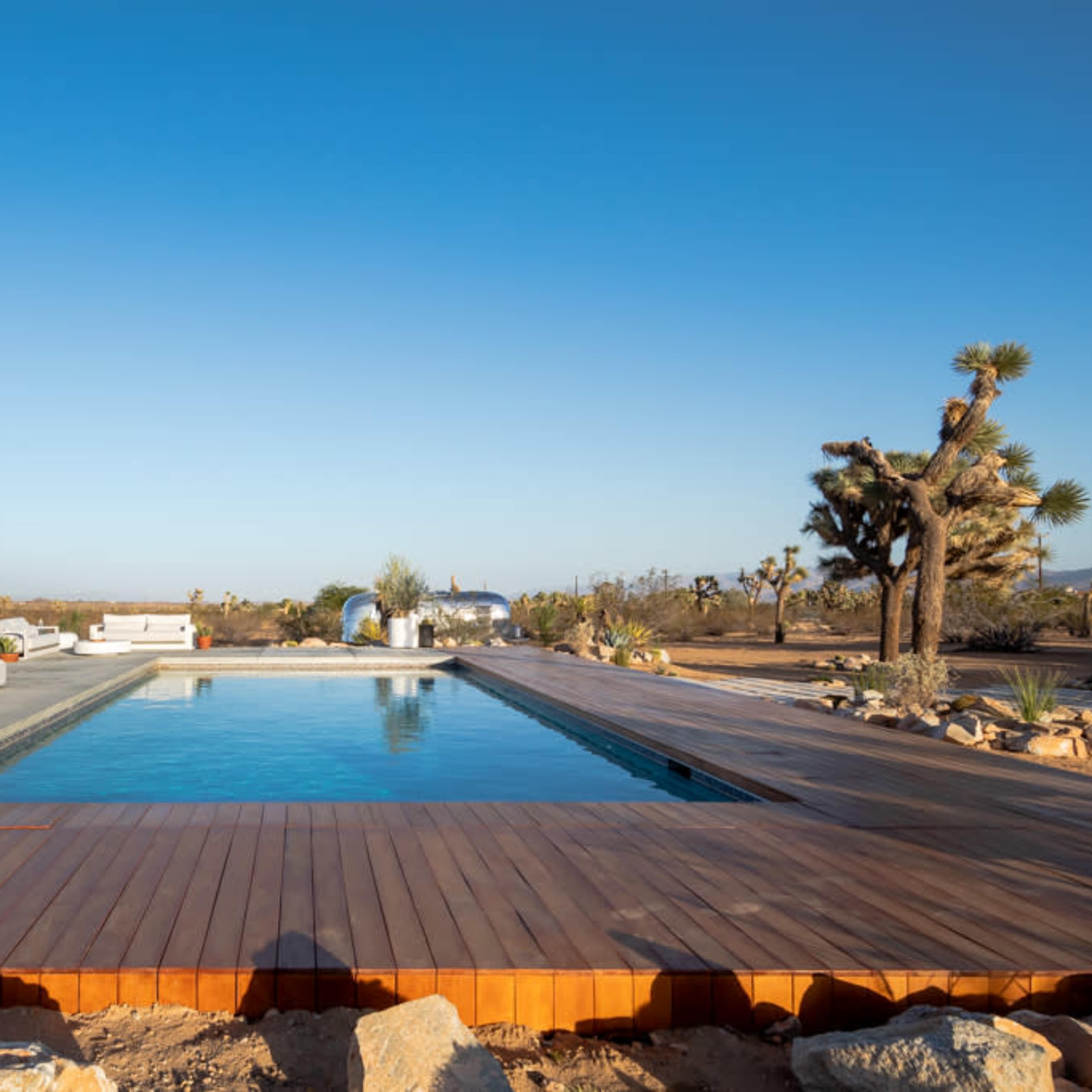 A swimming pool surrounded by a wooden deck and modern outdoor furniture in a desert landscape.