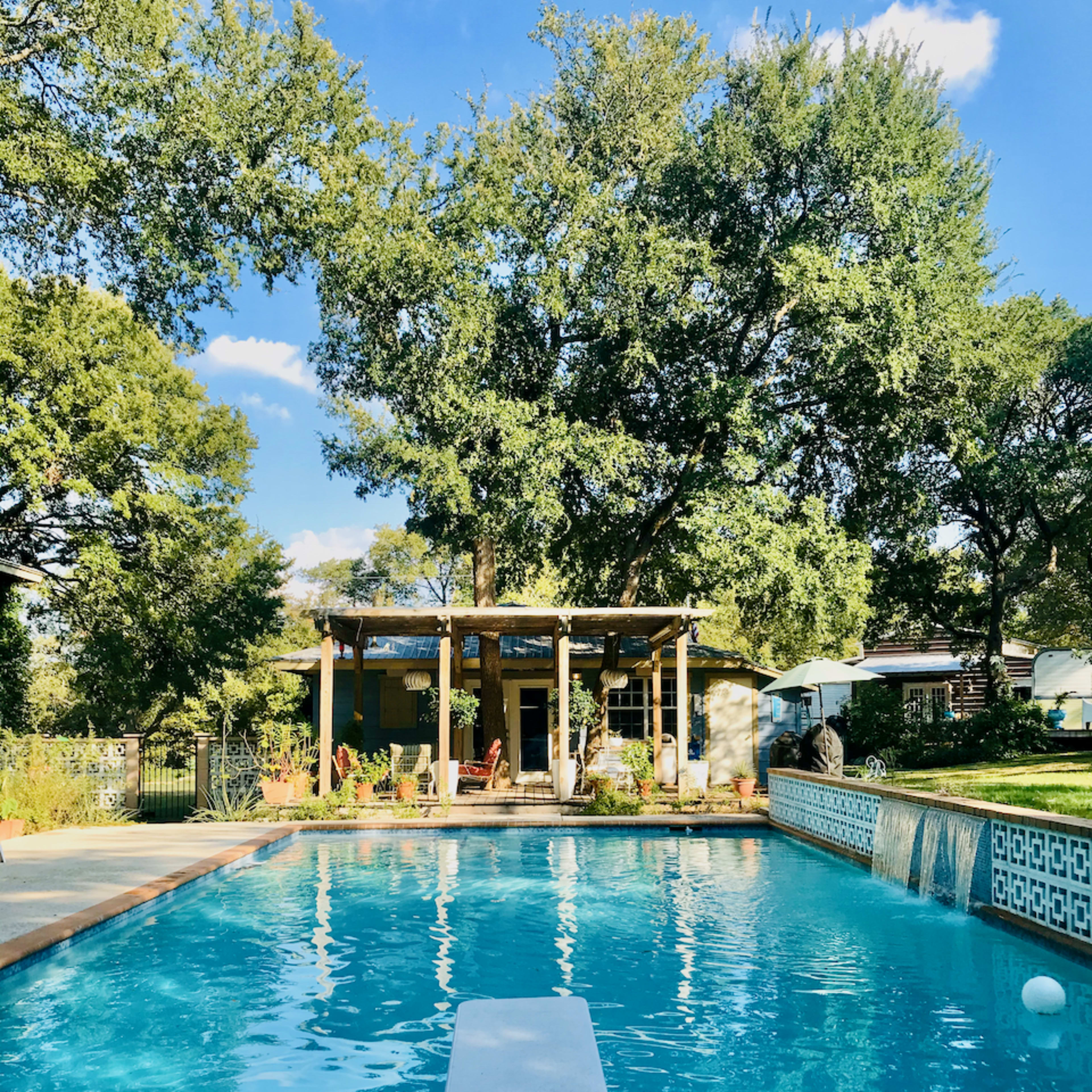 A clear swimming pool features a diving board and is surrounded by trees and a house with a shaded patio in the background.