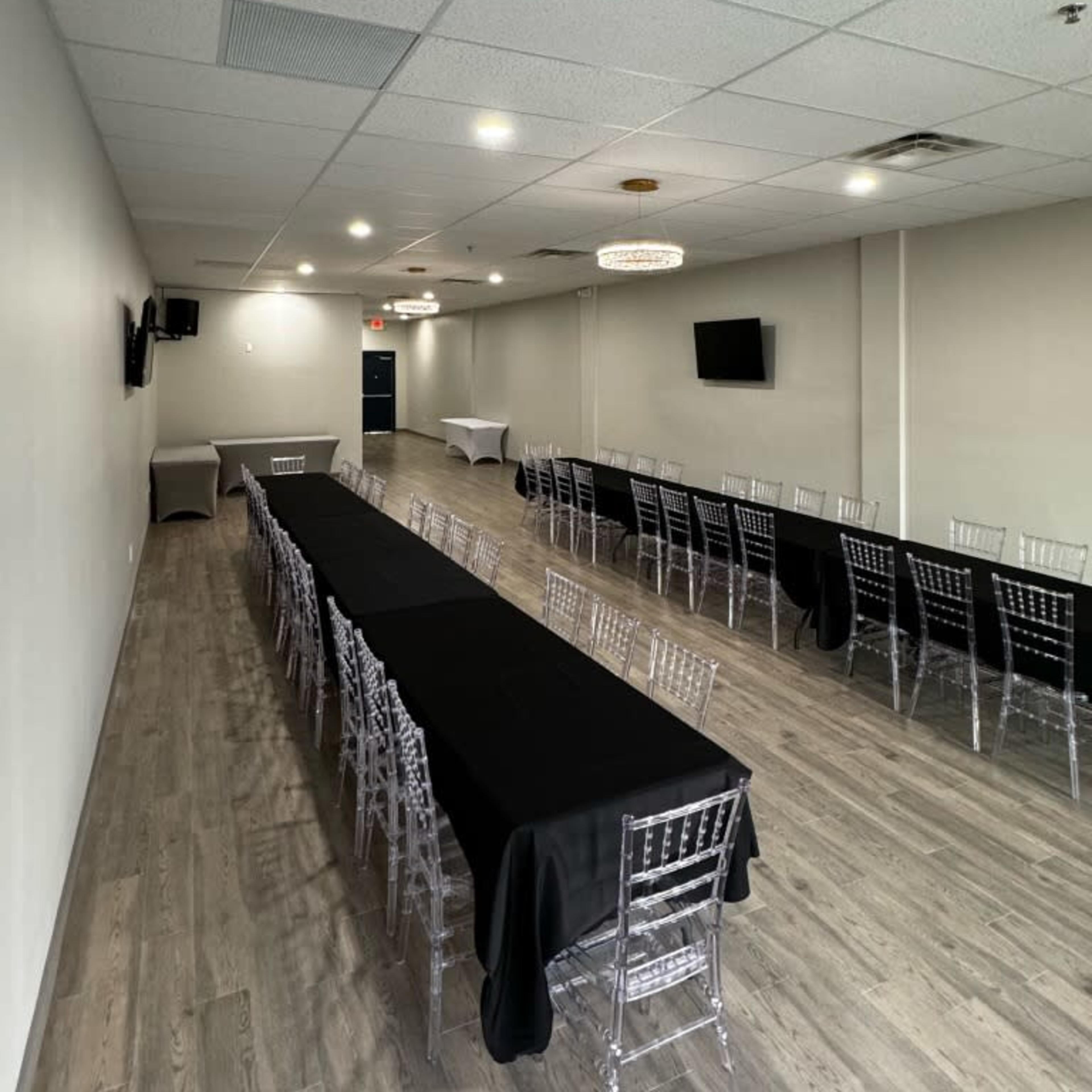 A spacious meeting room features long tables covered with black tablecloths and clear plastic chairs arranged in rows, with modern lighting overhead.