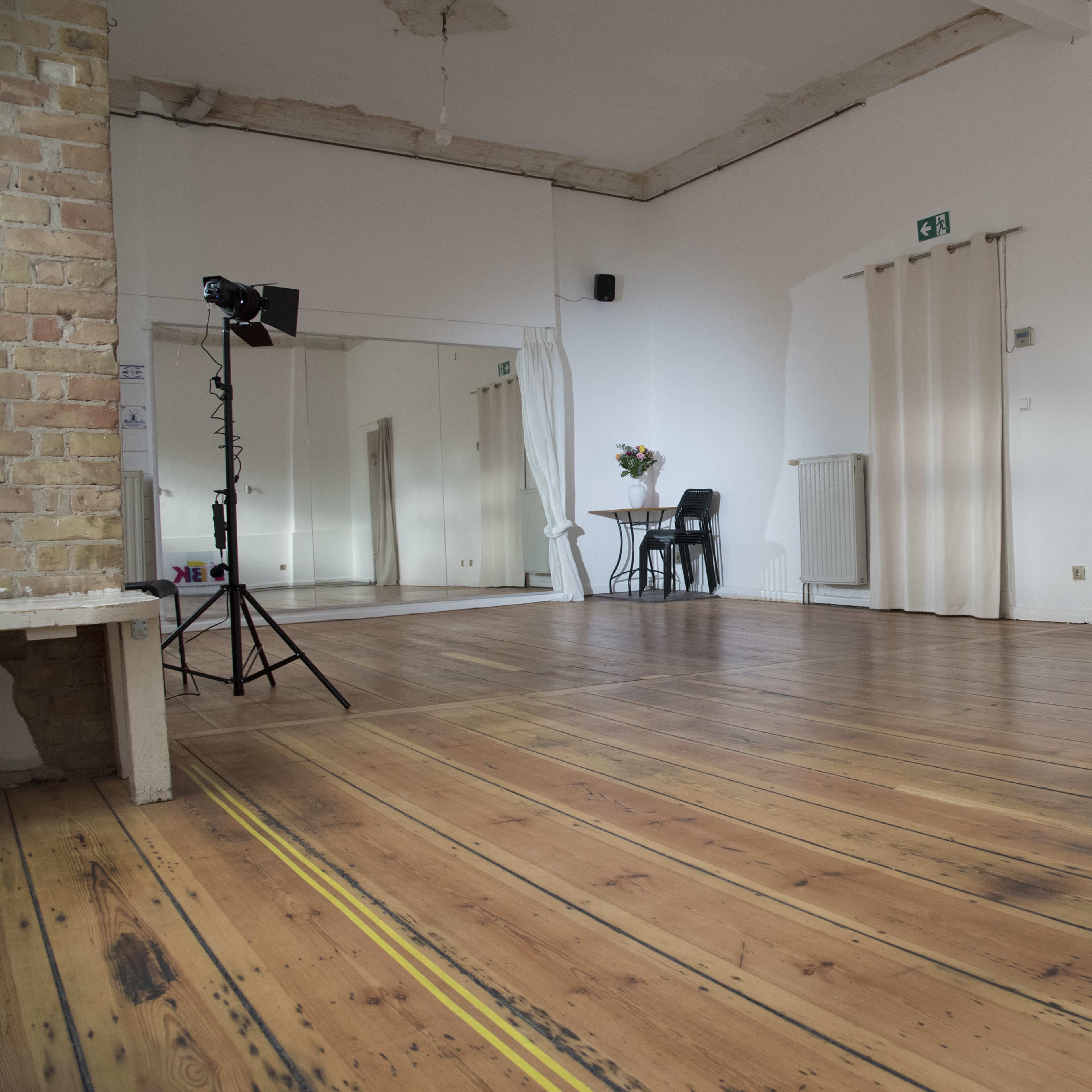 A spacious, empty room with wooden flooring, a standing light equipment, a chair, and partially closed curtains.