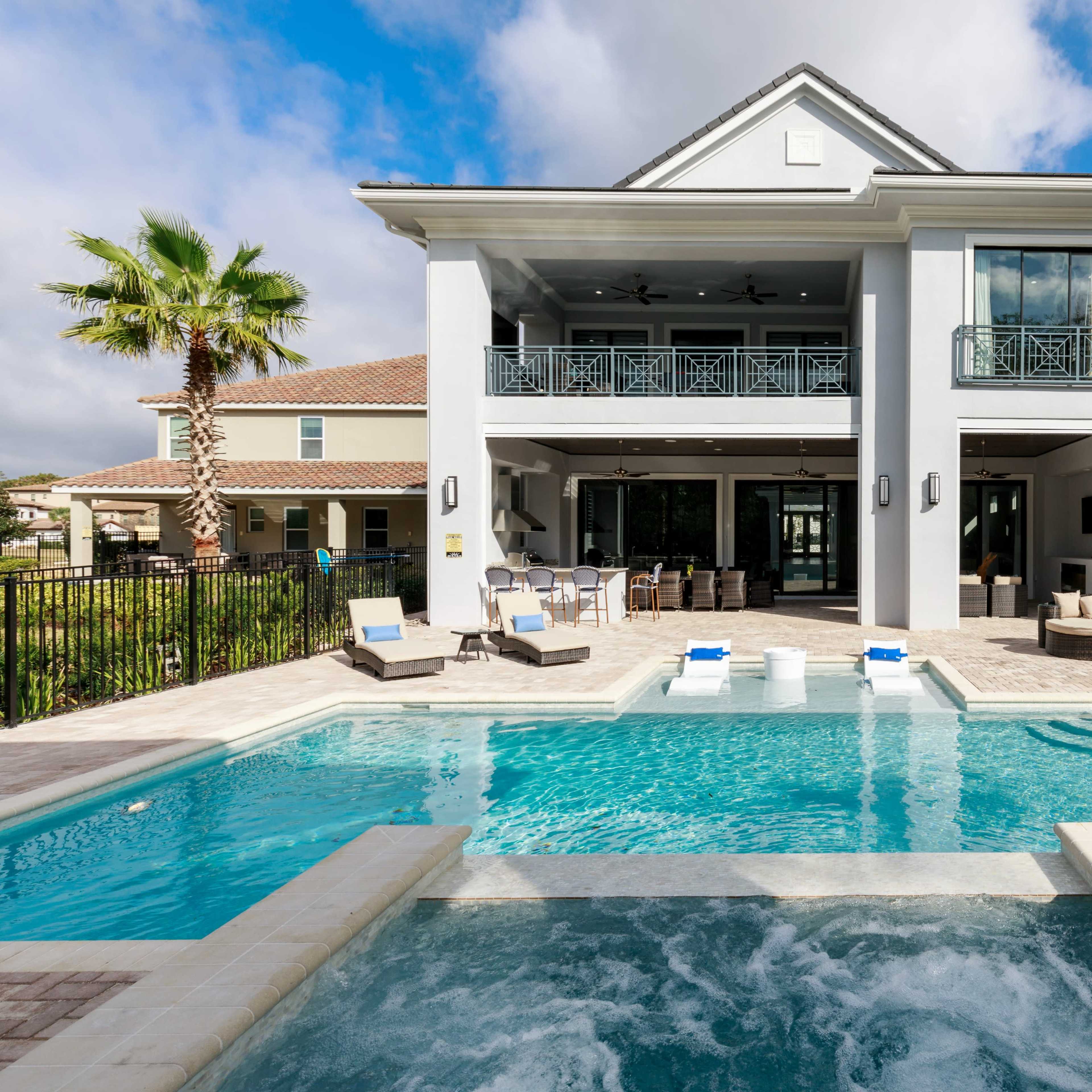 A modern house with a two-story structure, featuring a spacious pool area with a hot tub, surrounded by a fenced patio and palm trees.