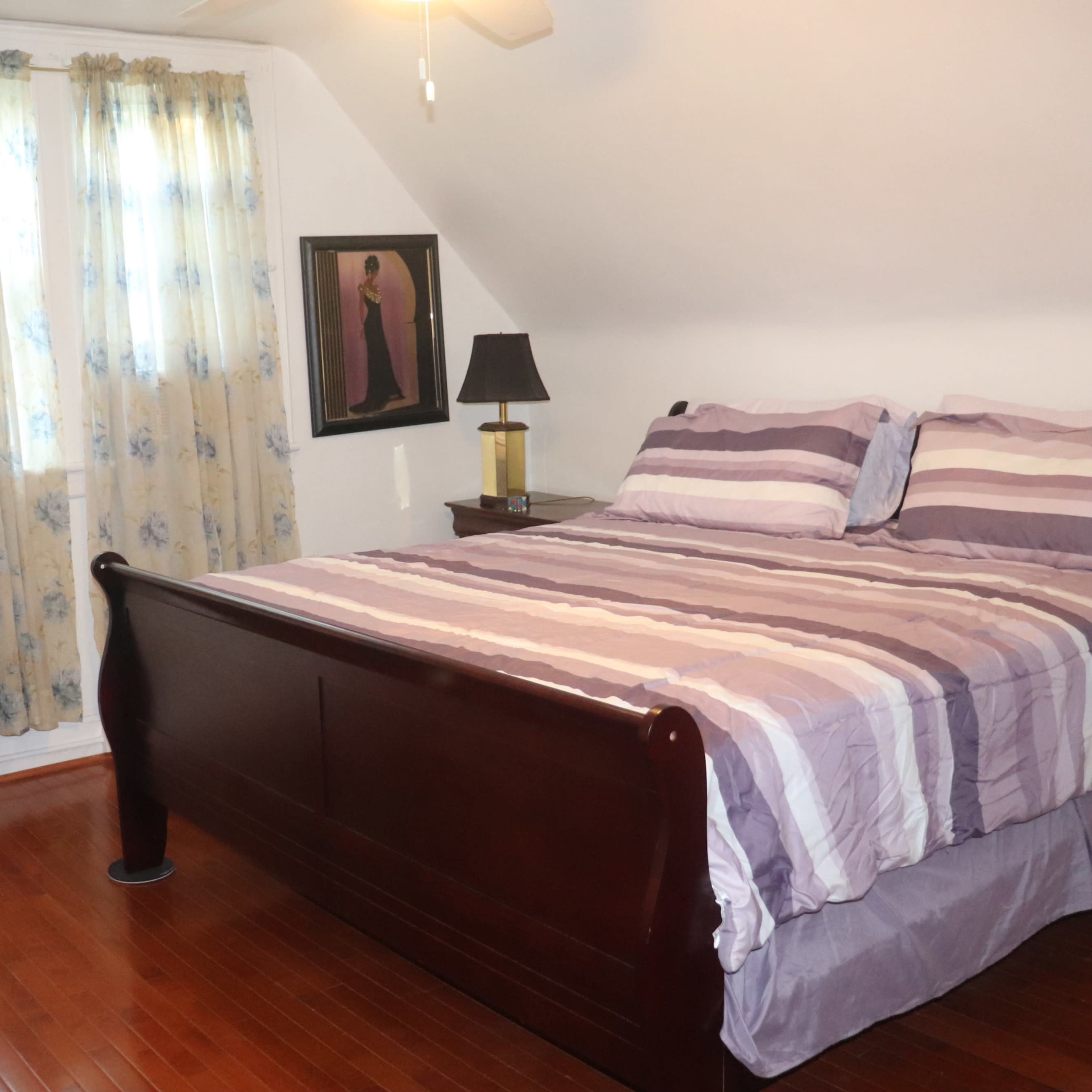 A bedroom with a wooden sleigh bed, striped bedding, a bedside lamp, and two windows covered with light curtains.