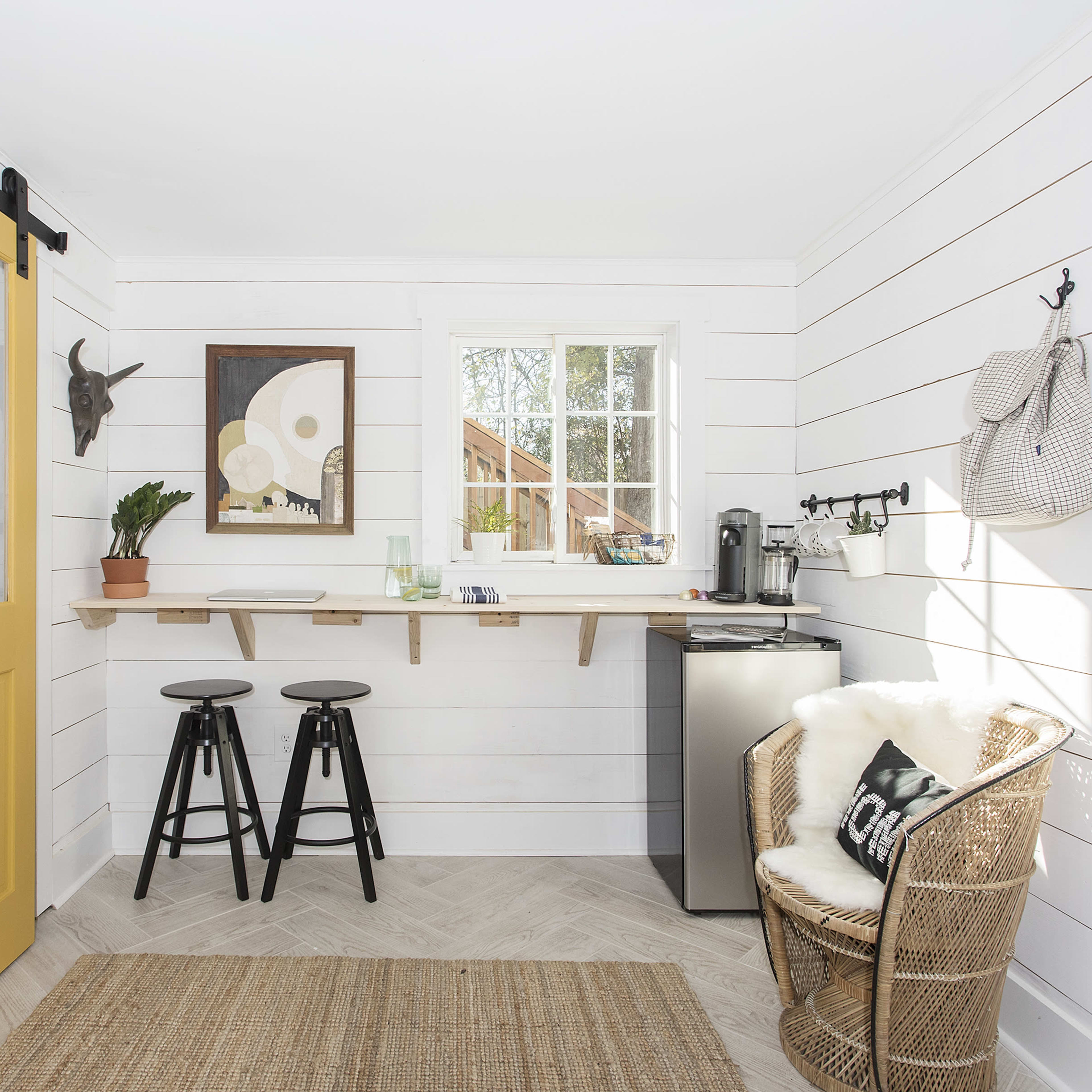 A small, bright room featuring a yellow sliding door, a wooden shelf, two black stools, a kitchenette with a mini fridge, and a rattan chair.