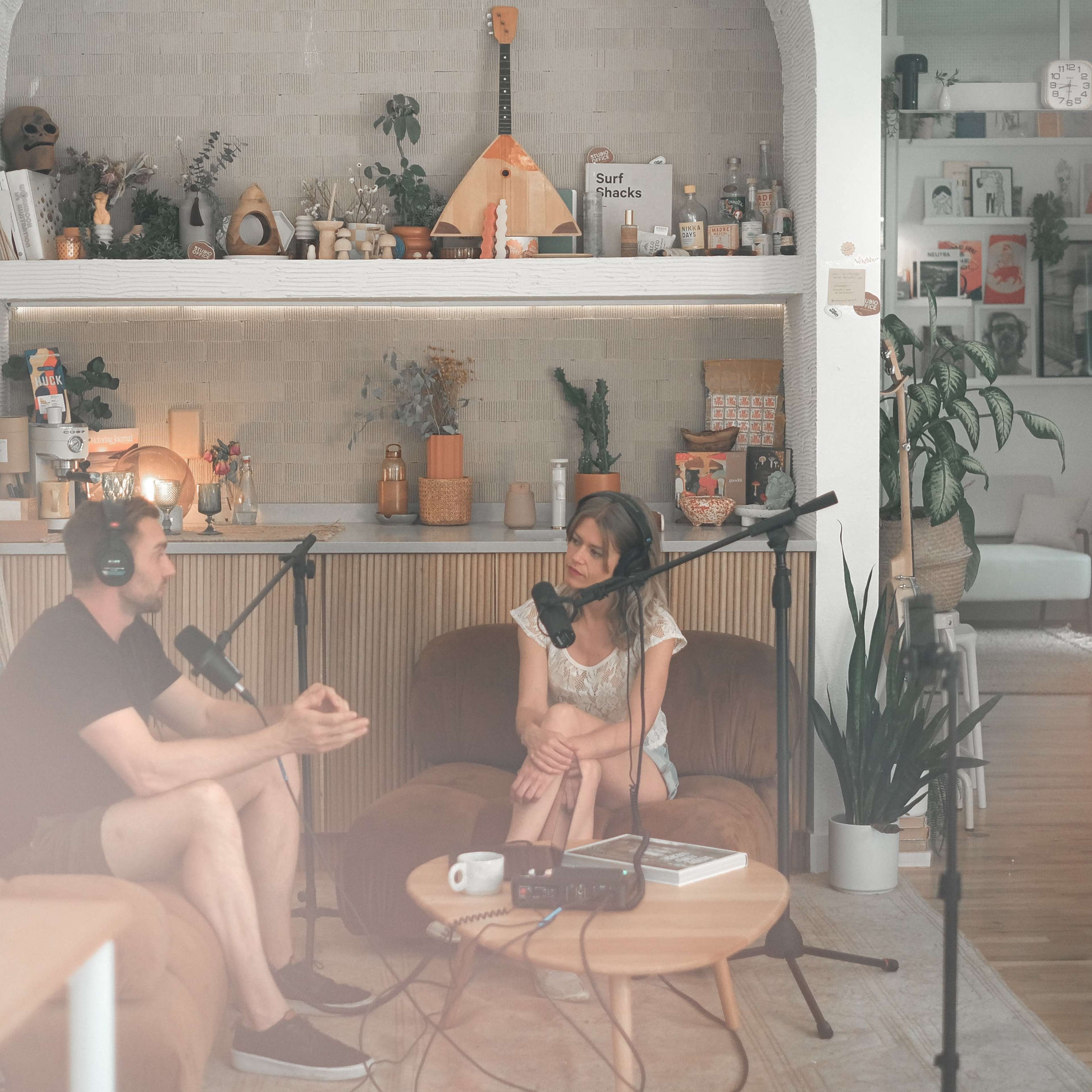 A man and a woman are engaged in a conversation in a cozy, well-decorated podcast studio with microphones and cameras set up.