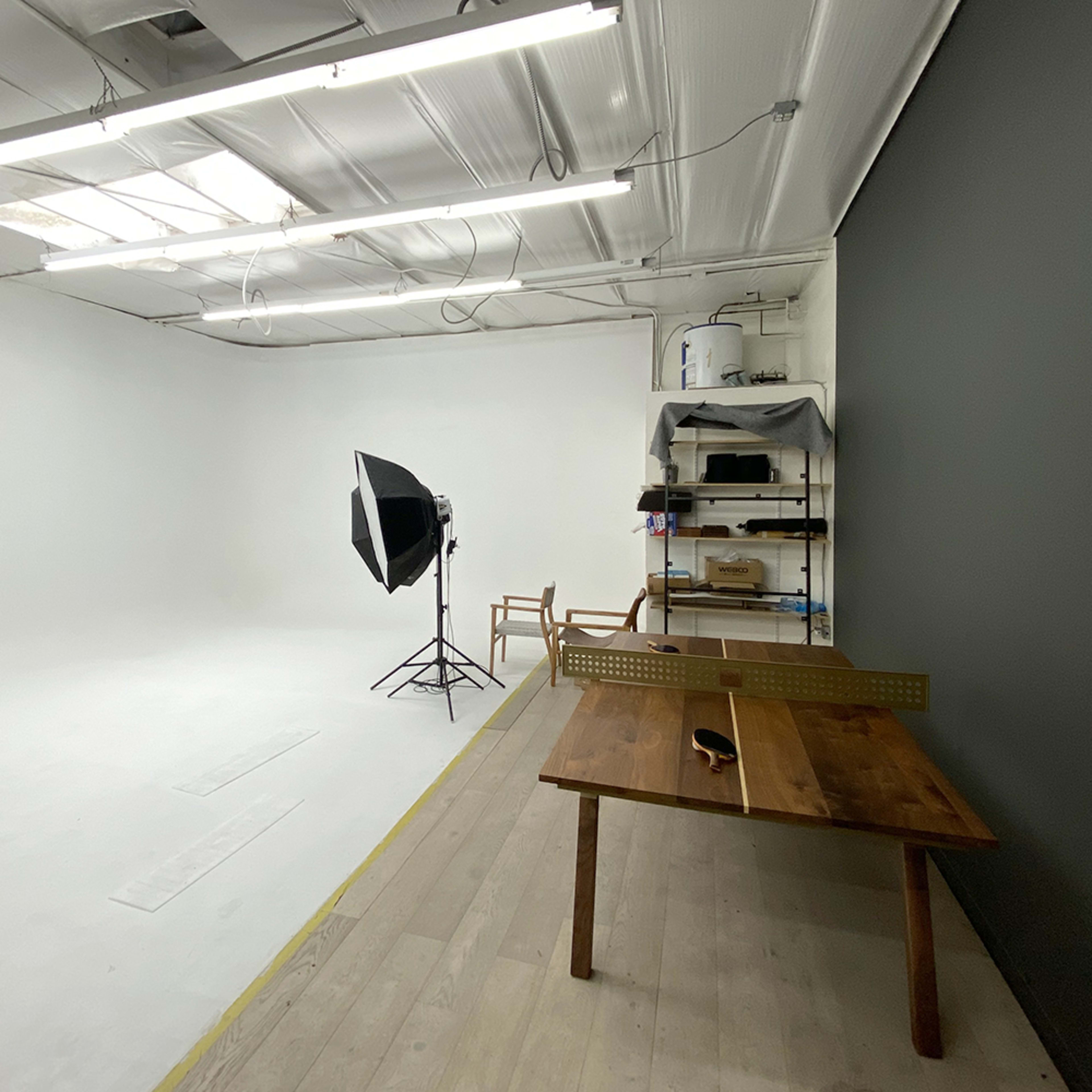 A bare studio space with white walls, a wooden table, a chair, and a large light source on a stand.