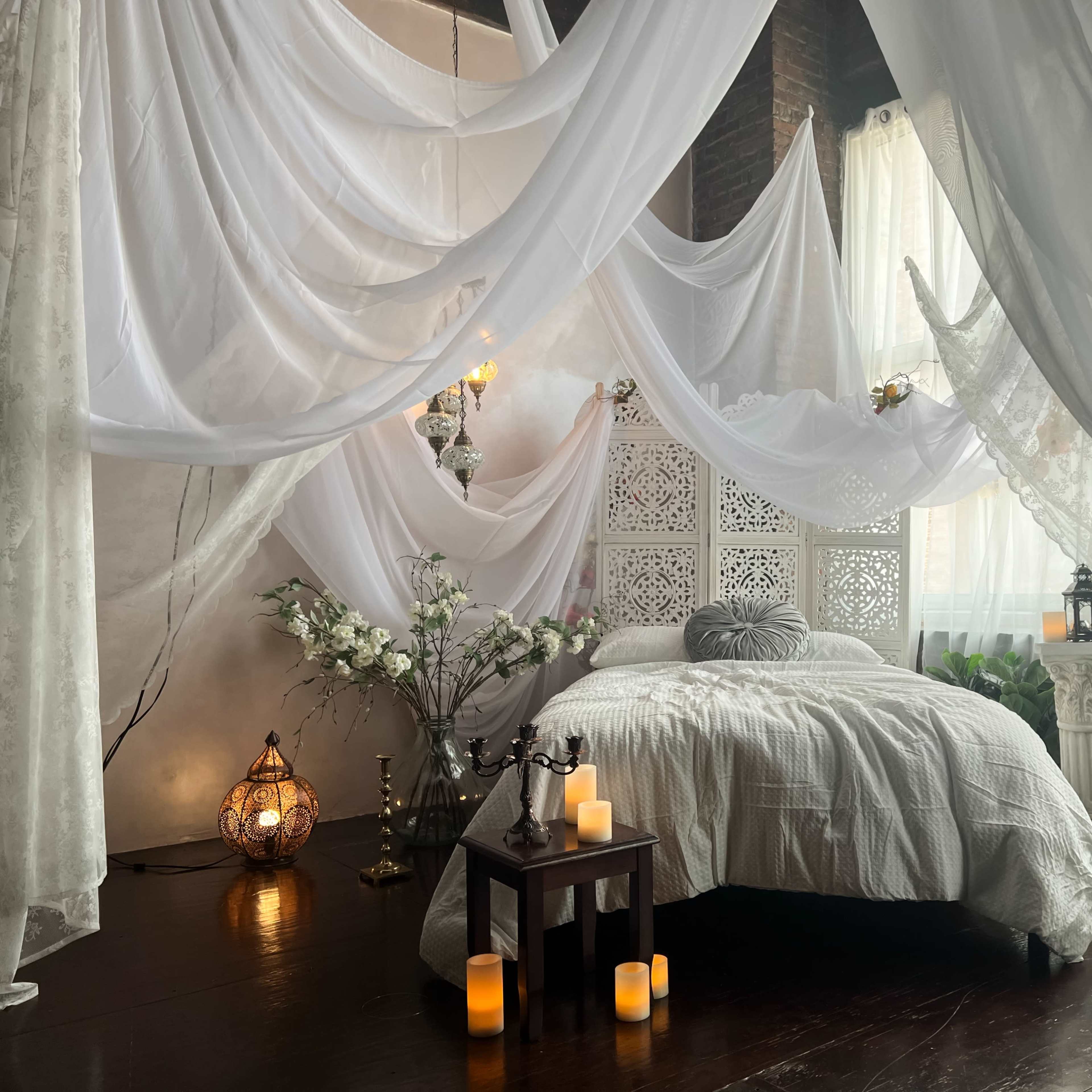 A serene bedroom adorned with flowing white drapes and a neatly made bed surrounded by decorative elements like candles and floral arrangements.