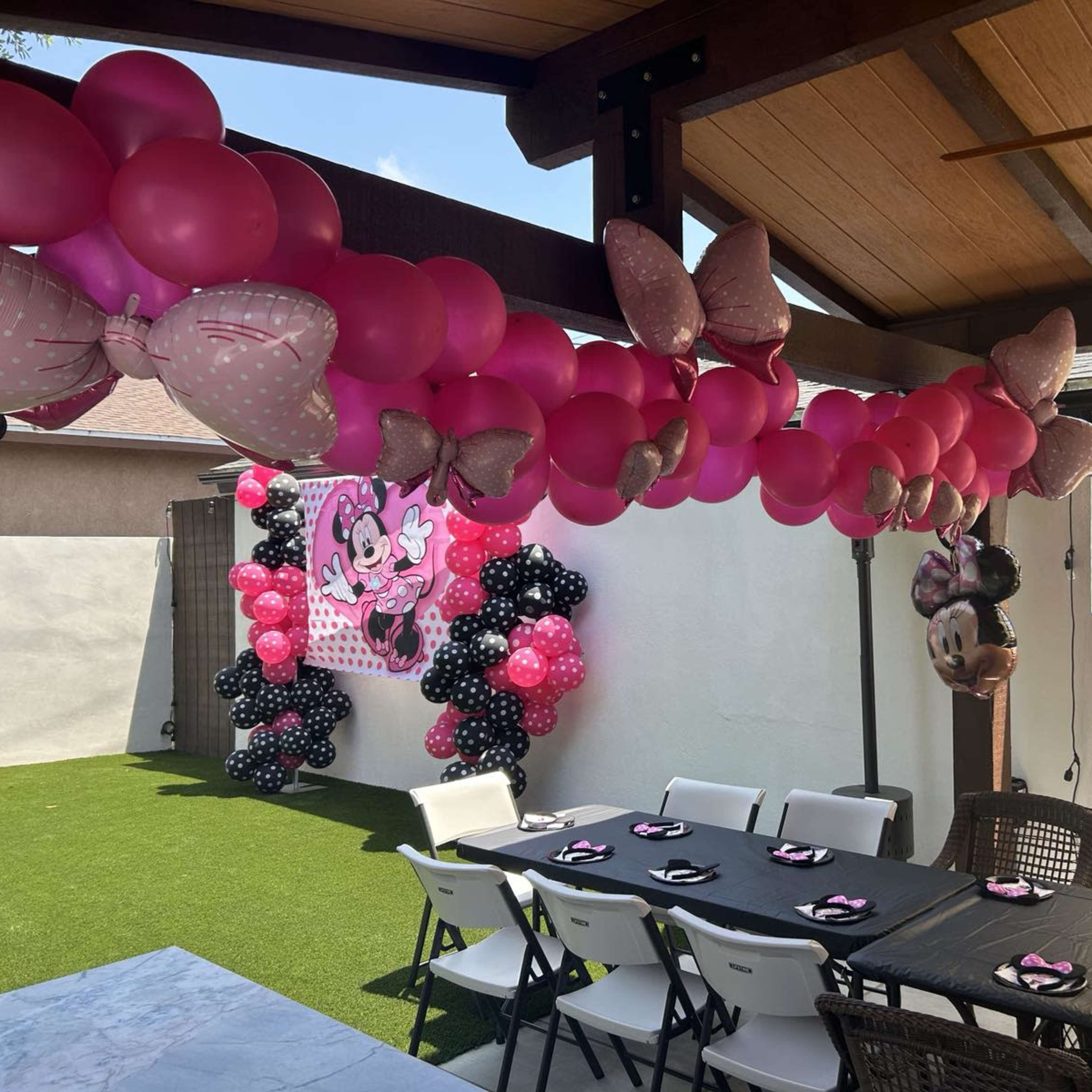 A decorated outdoor party space featuring pink and black balloons, including Minnie Mouse-themed elements, with a long table set for guests.