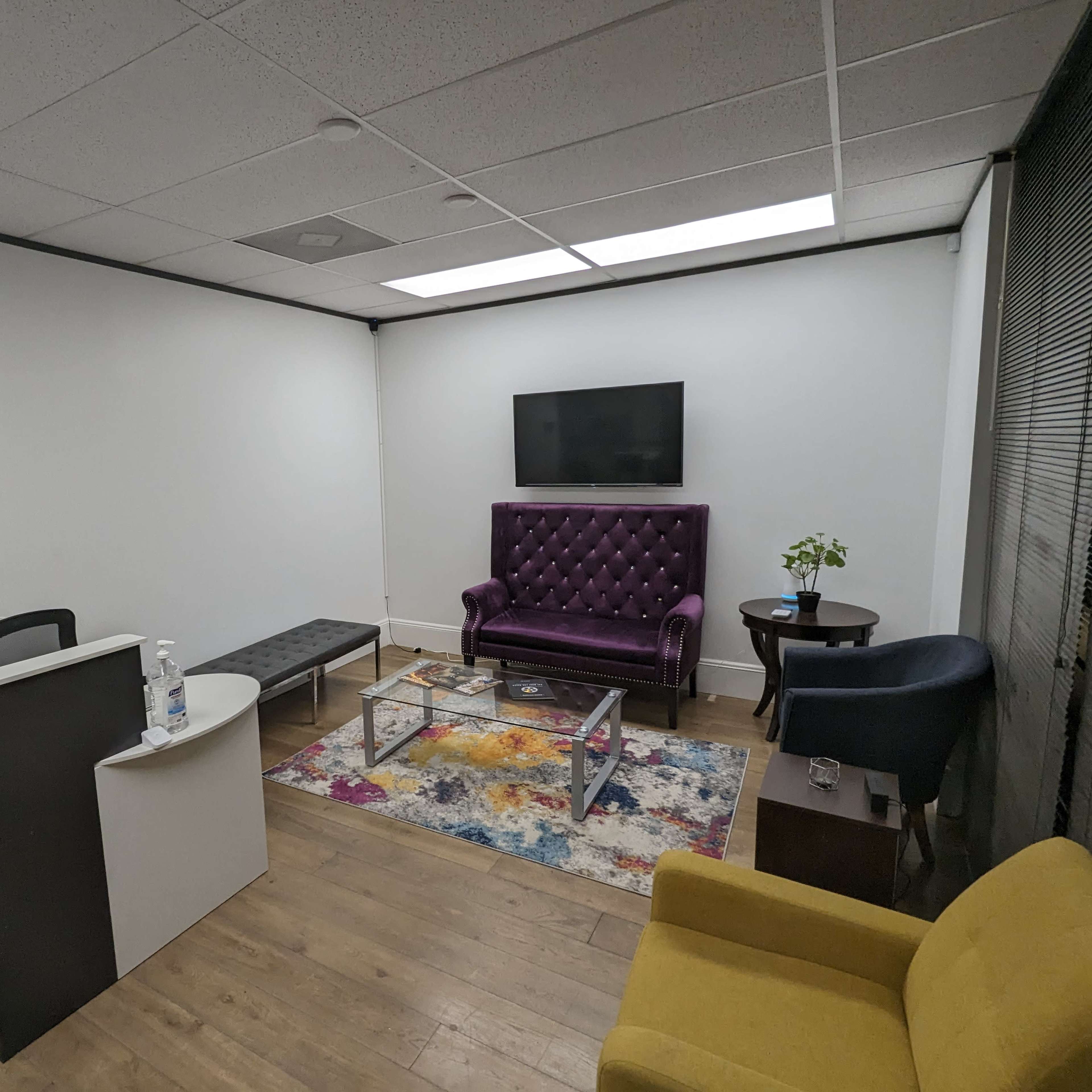 A modern office space with a purple upholstered sofa, a glass coffee table, a small plant on a side table, and a reception area featuring a minimalist desk.