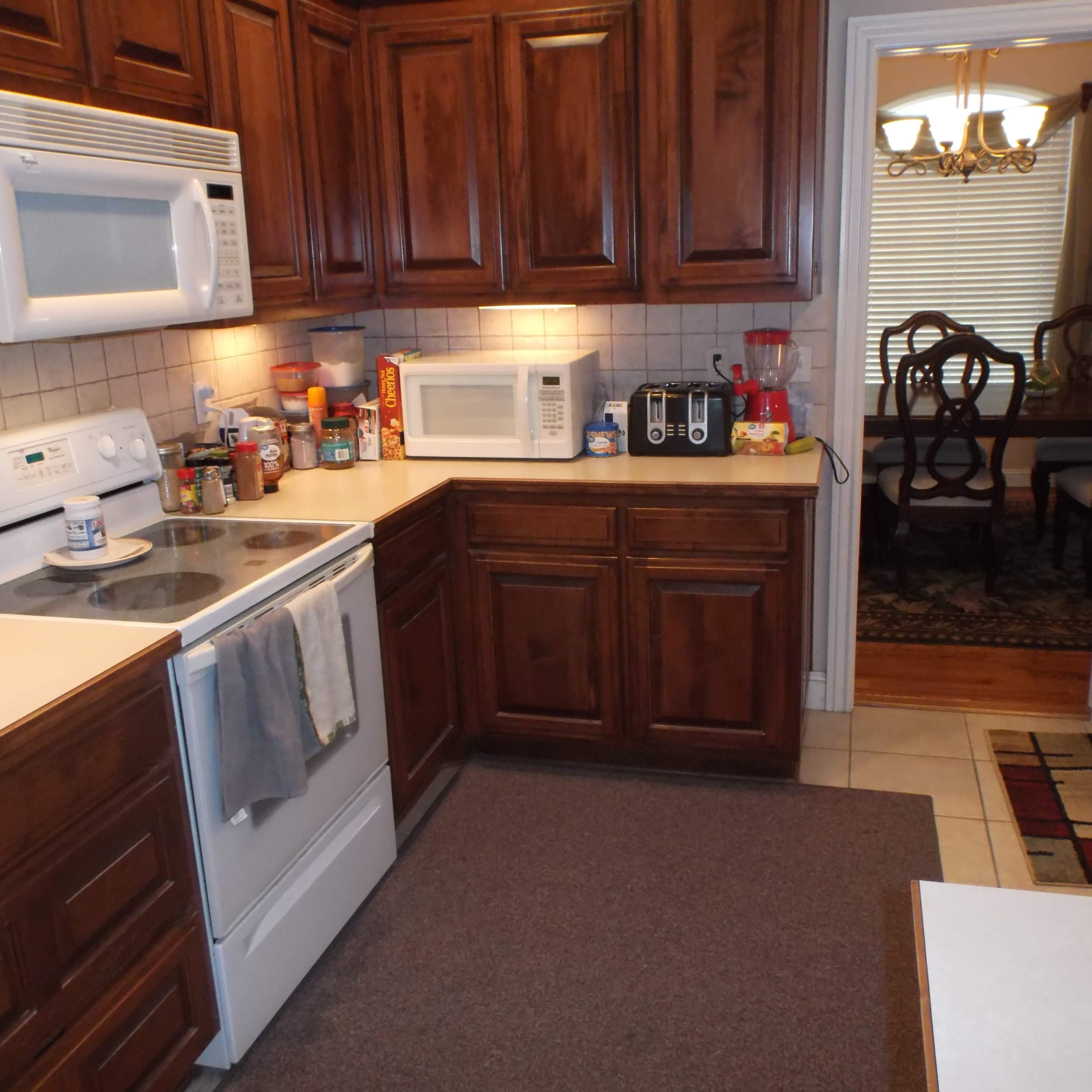 A kitchen with wooden cabinets, a stove, microwave, and various appliances on the countertops, leading to a dining area visible in the background.