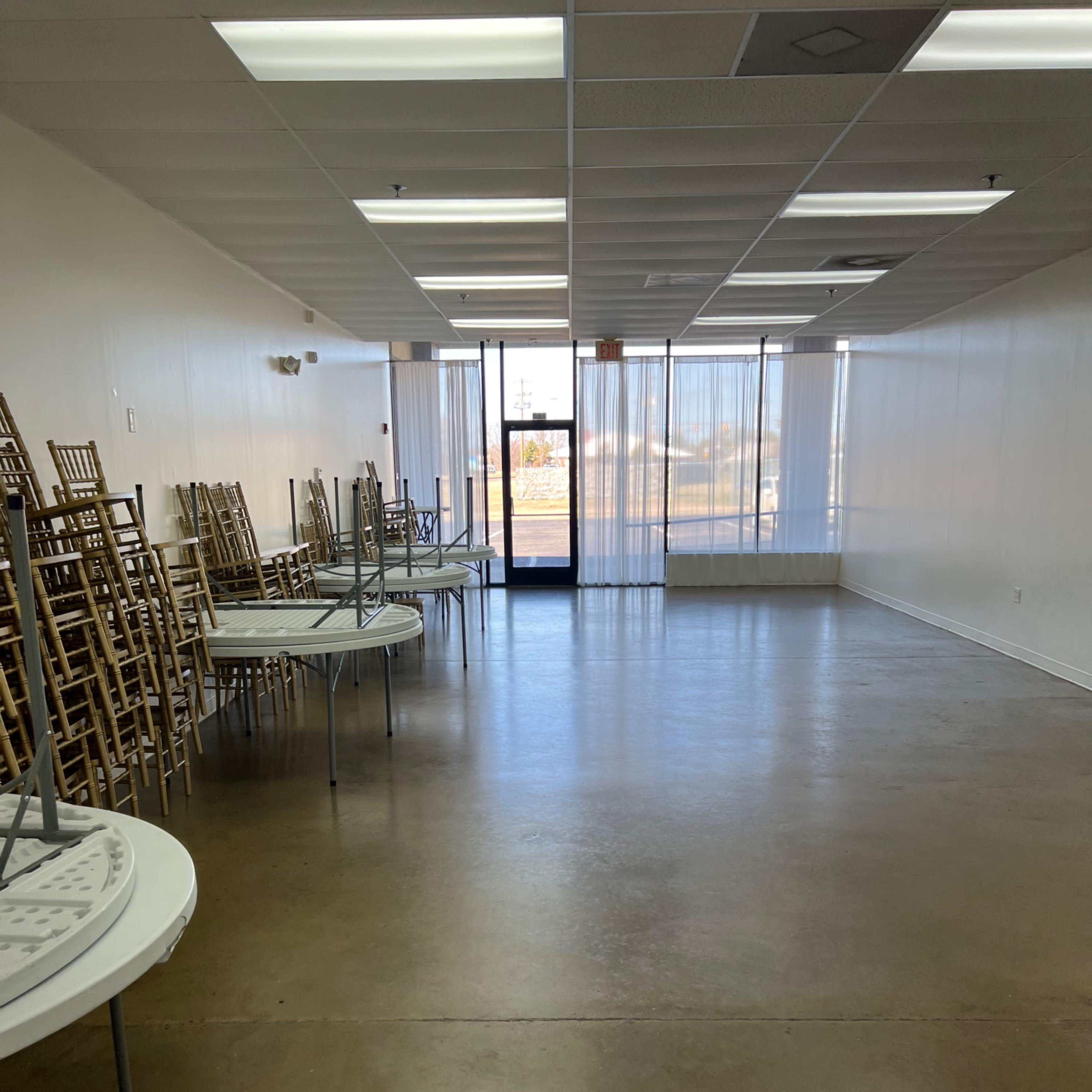 A spacious, empty room with stacked chairs against the wall and tables turned upside down on the floor.