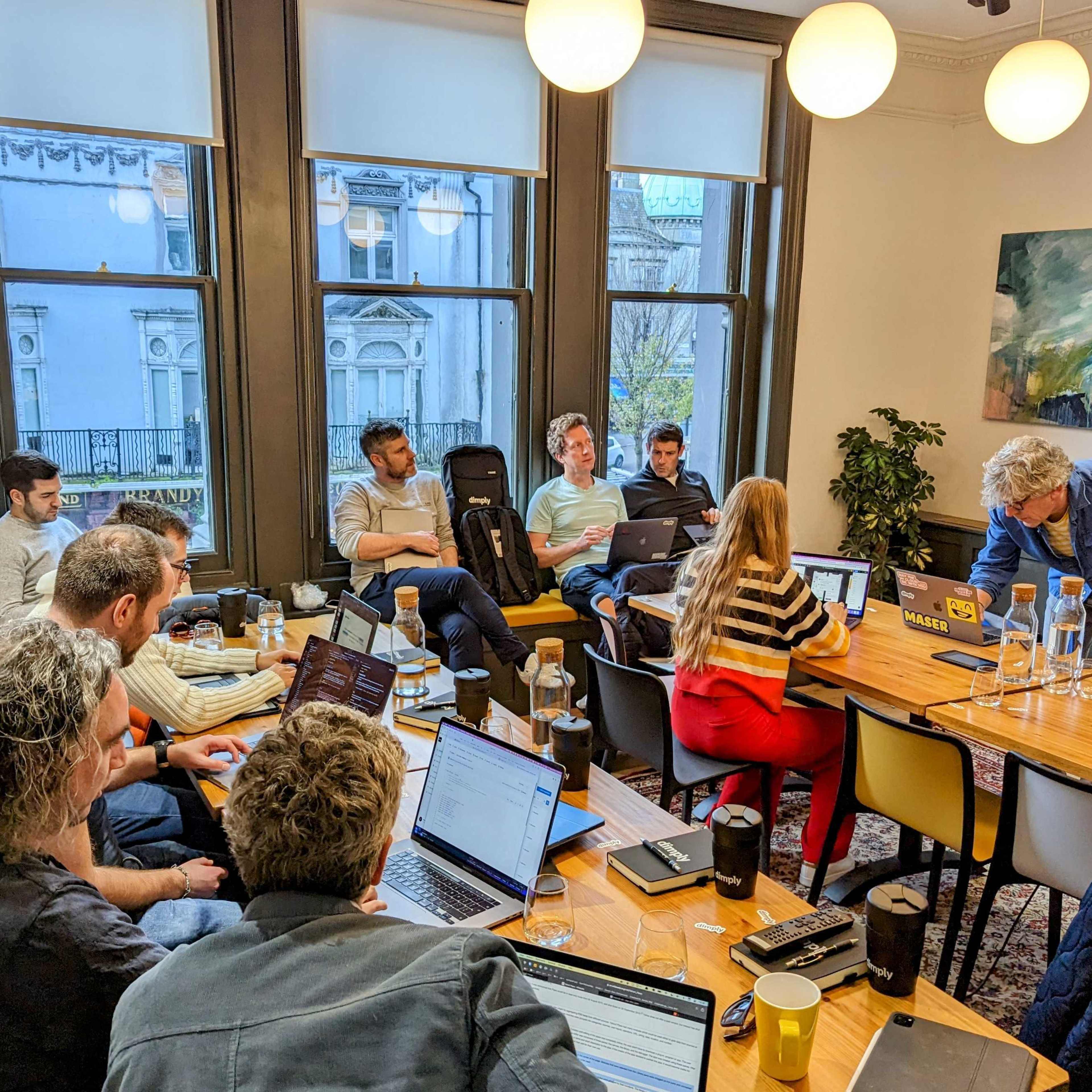 A group of people is seated around a wooden table in a meeting room, with laptops open and a presentation displayed on a screen.