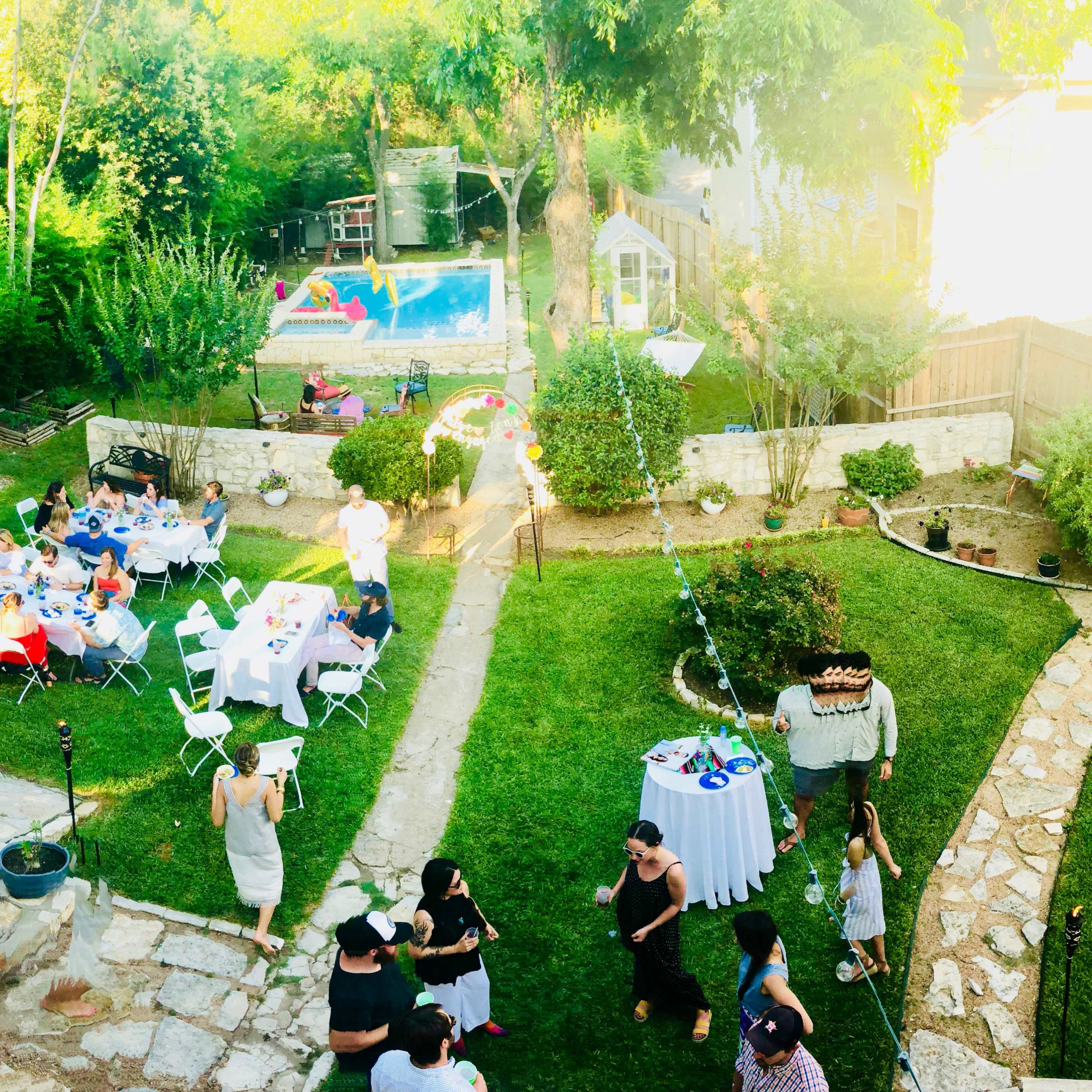 A backyard party is taking place with tables set for dining, guests socializing, and a swimming pool visible in the background.