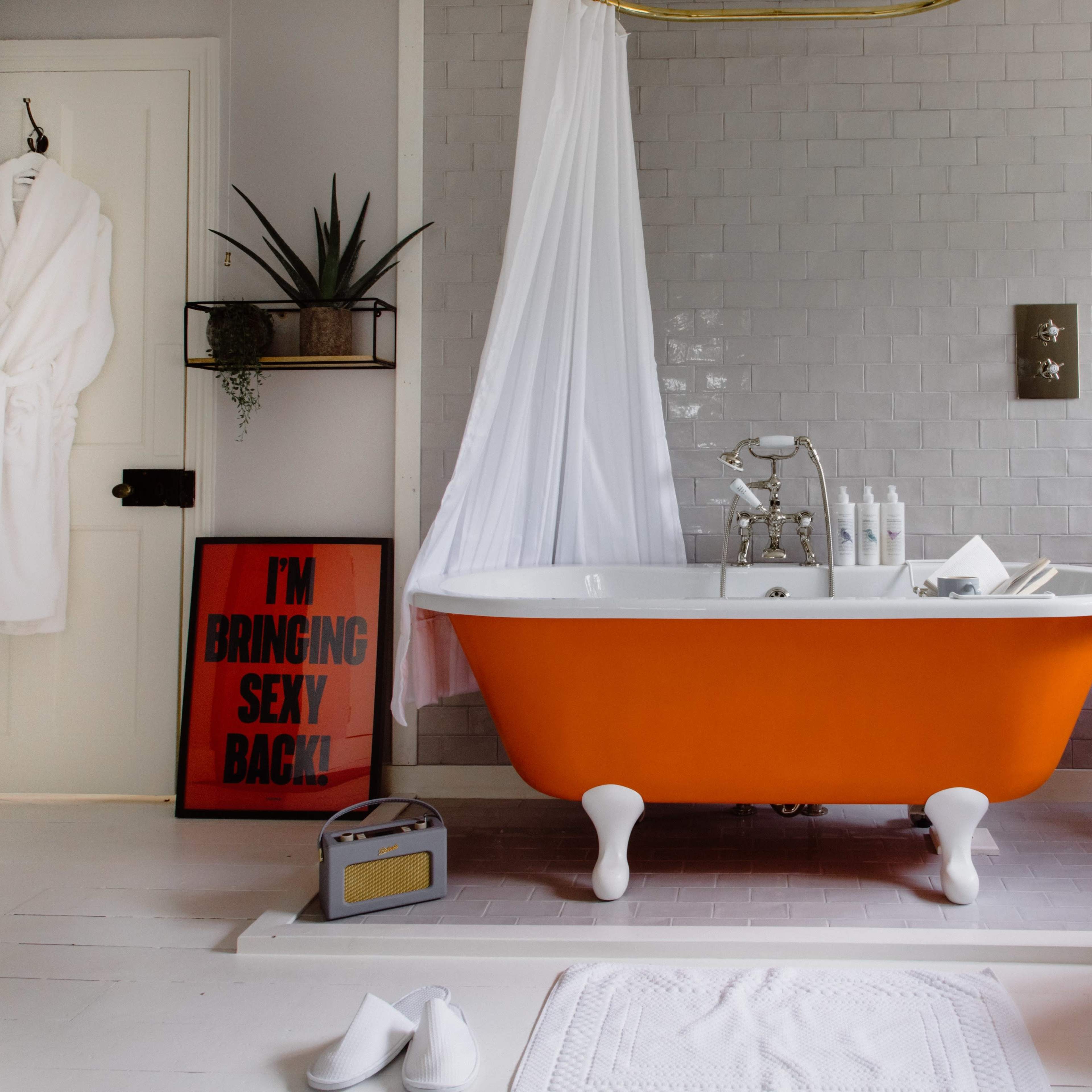 The bathroom features a vintage orange bathtub with a metal faucet, surrounded by a black radiator, a wooden chair, and decorative plants.