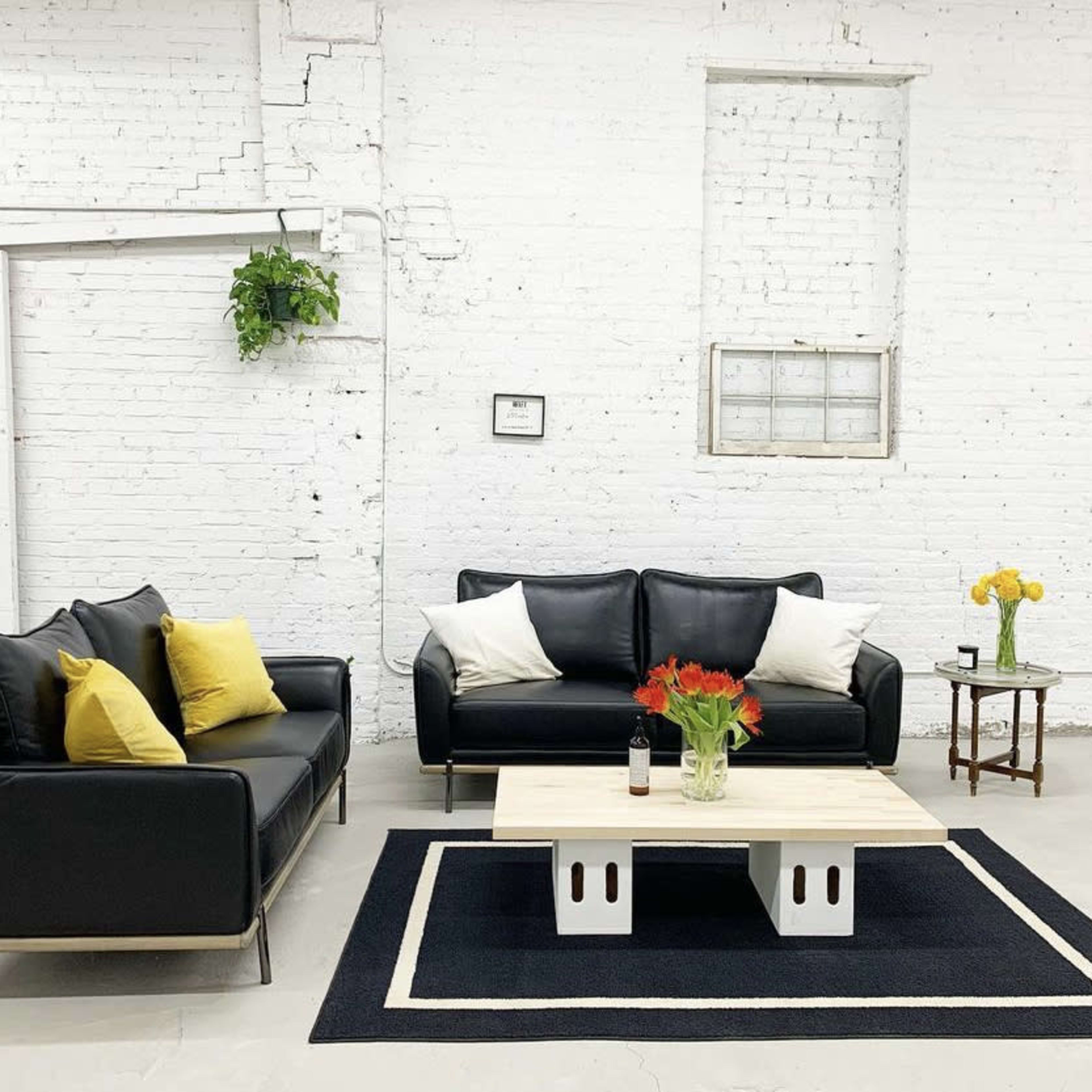 A minimalistic living room with a black sofa, a loveseat with yellow cushions, a wooden coffee table, and a small side table, all set against a white brick wall.