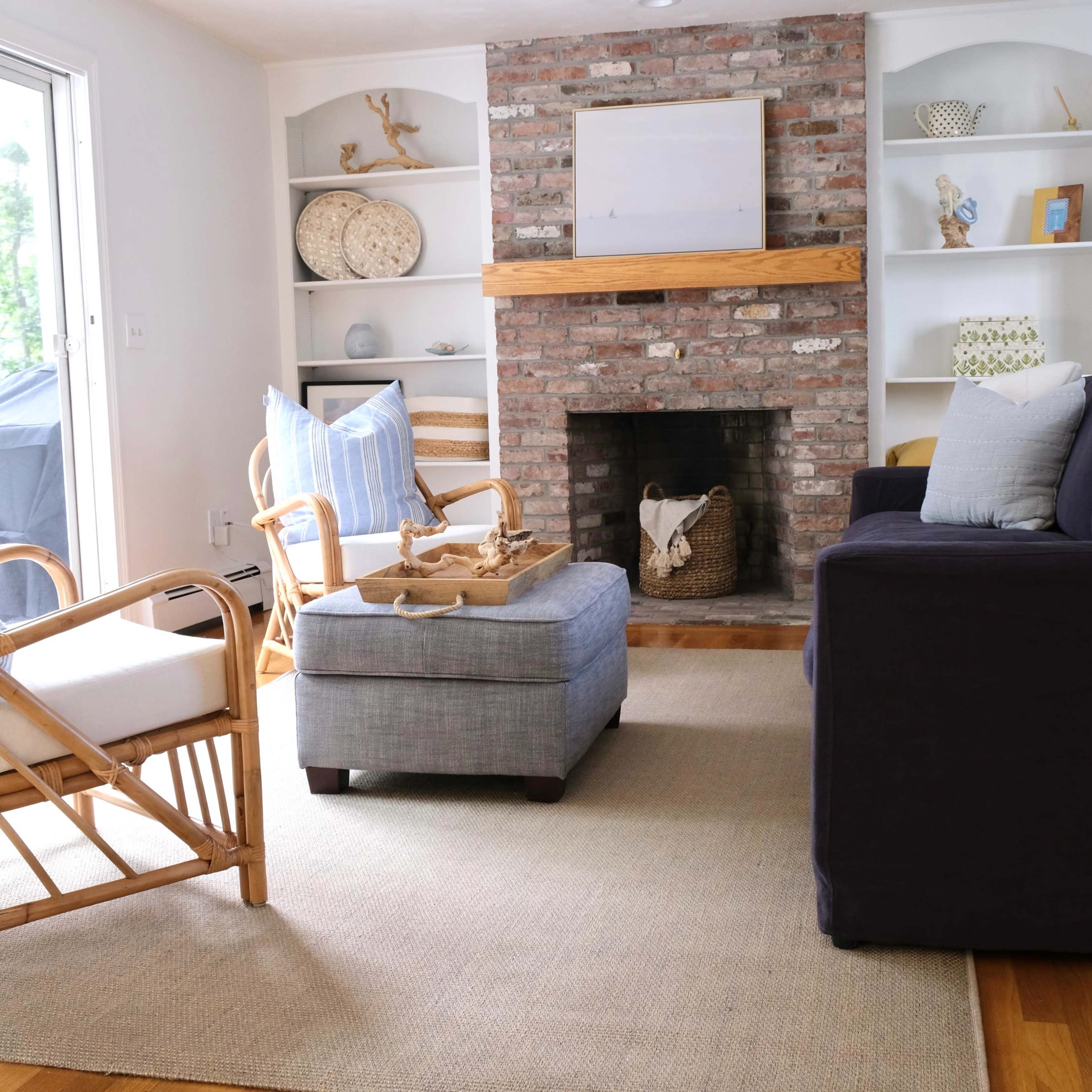 A cozy living room with a brick fireplace, a blue upholstered chair, a light gray ottoman, and two rattan chairs arranged around a neutral-colored area rug.