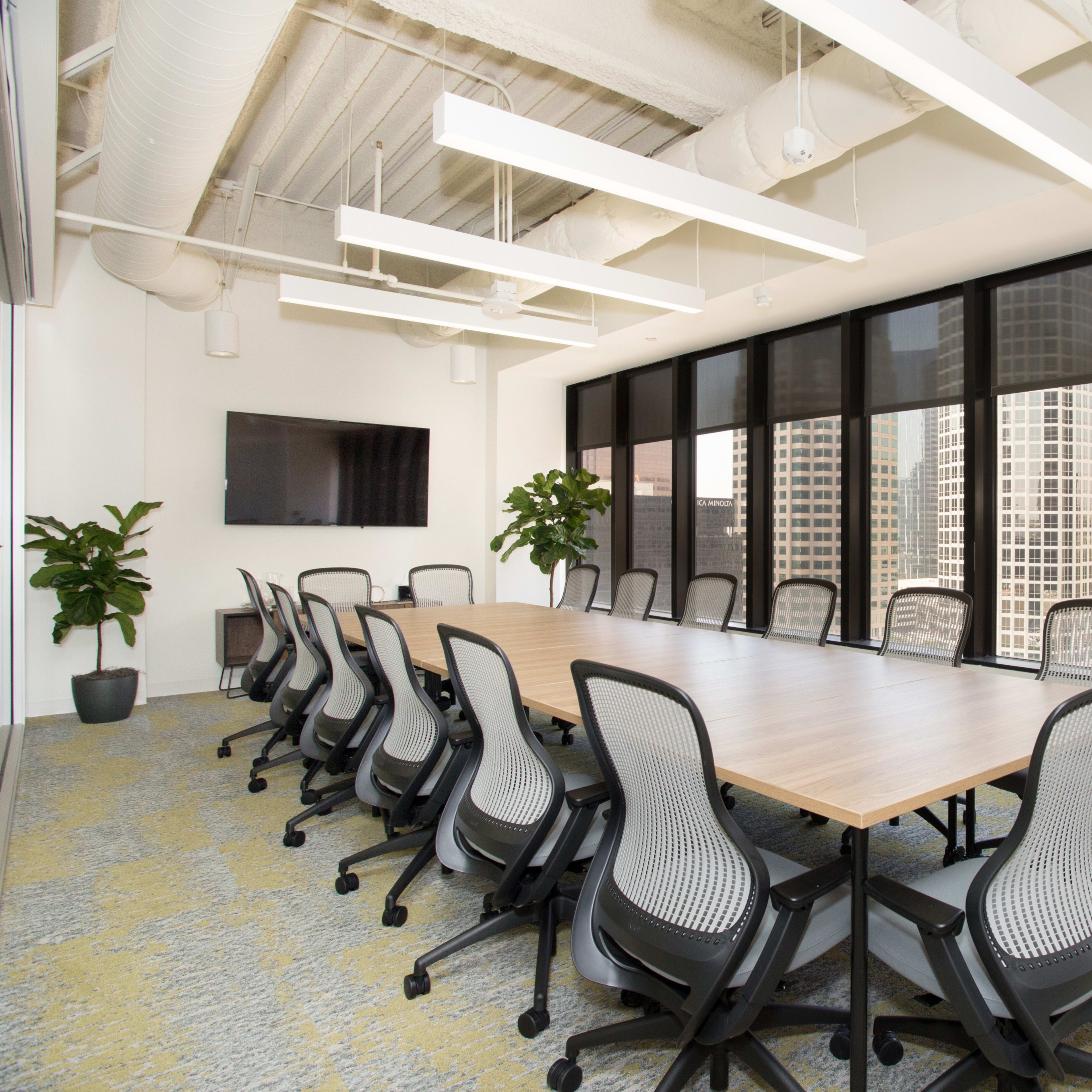 A conference room features a long wooden table surrounded by ergonomic chairs, with large windows offering a view of a city skyline.