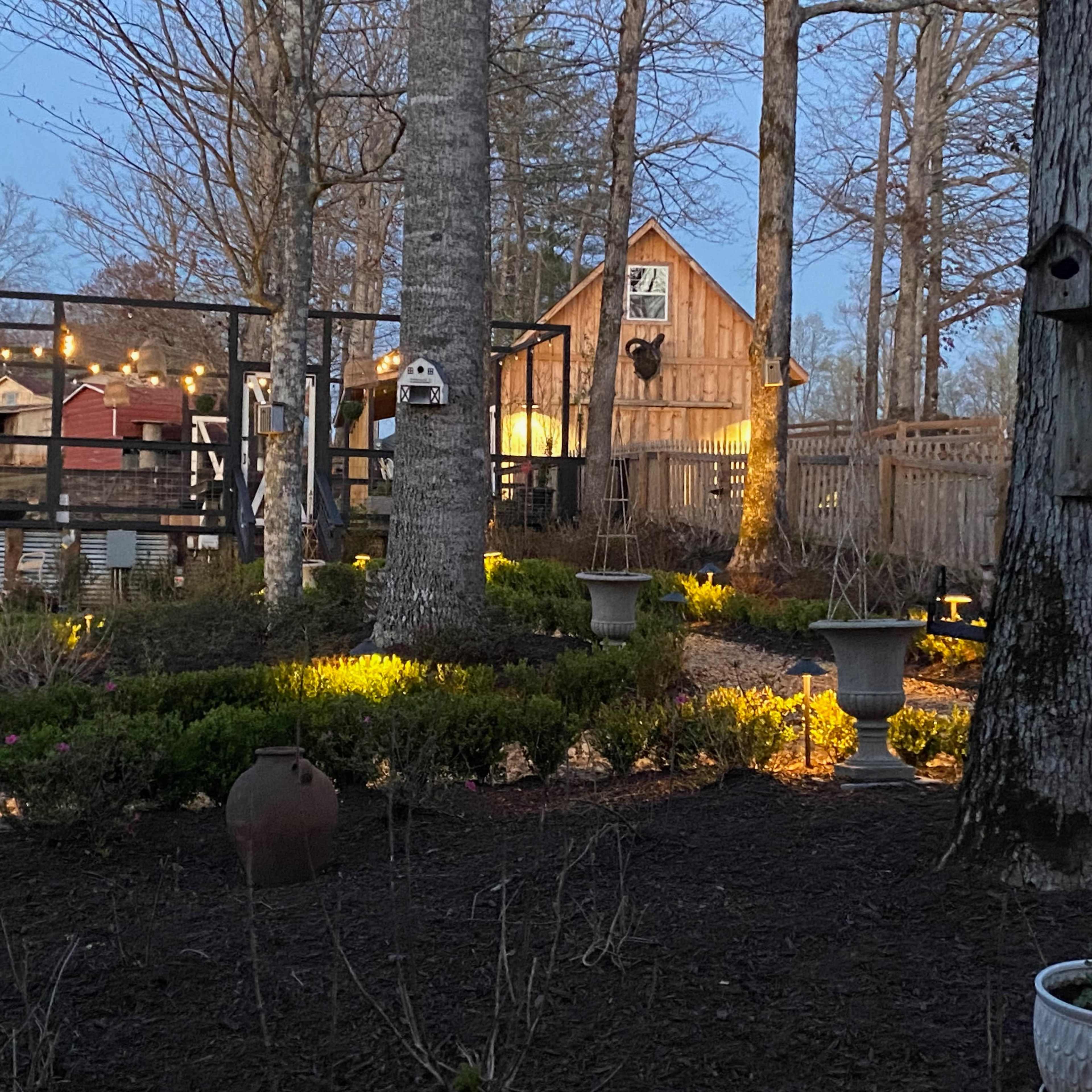 A landscaped garden area featuring stone paths, birdhouses, and a wooden cabin in the background, illuminated by string lights among the trees.