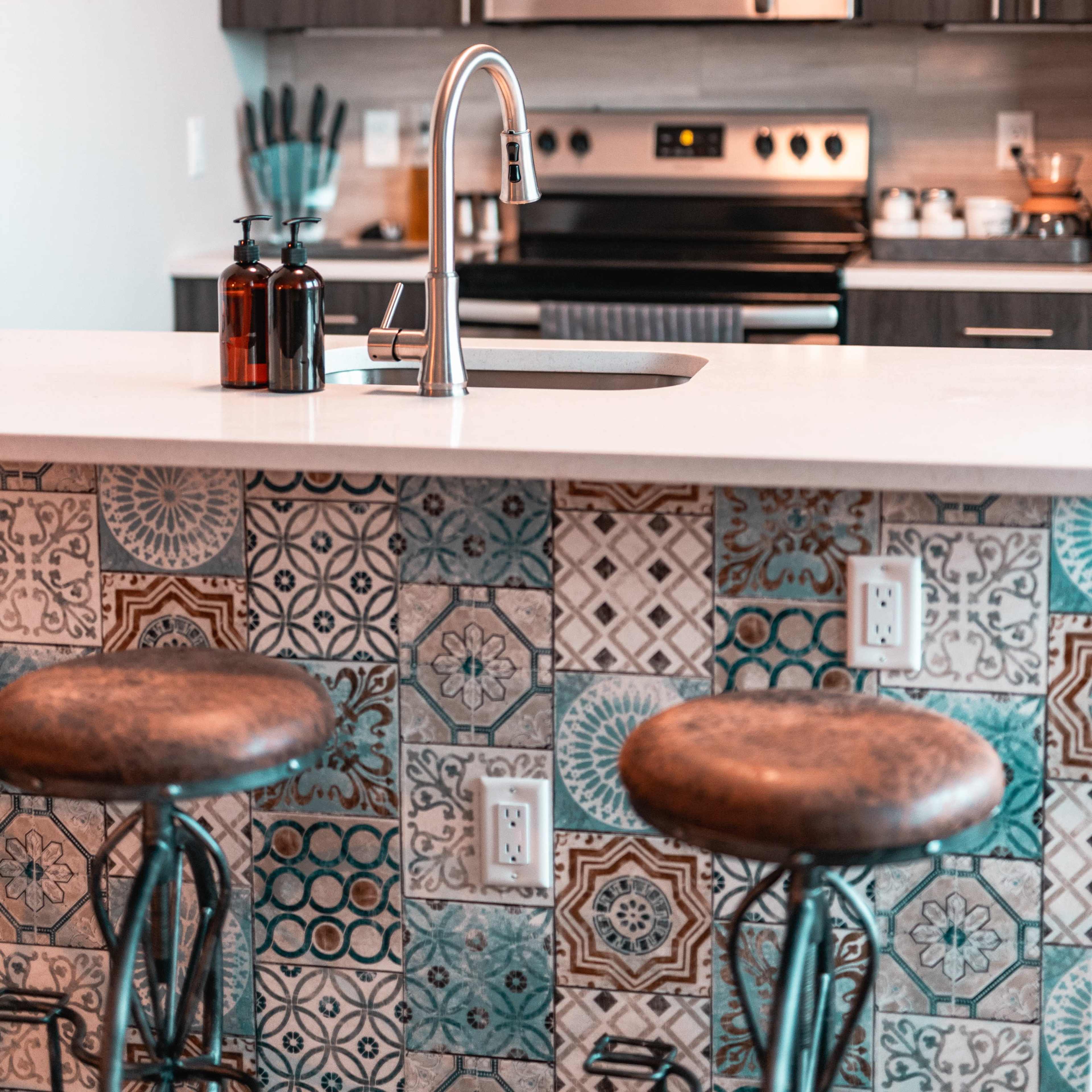 A modern kitchen with a countertop, a stainless steel sink, bar stools, and a decorative tiled backsplash.