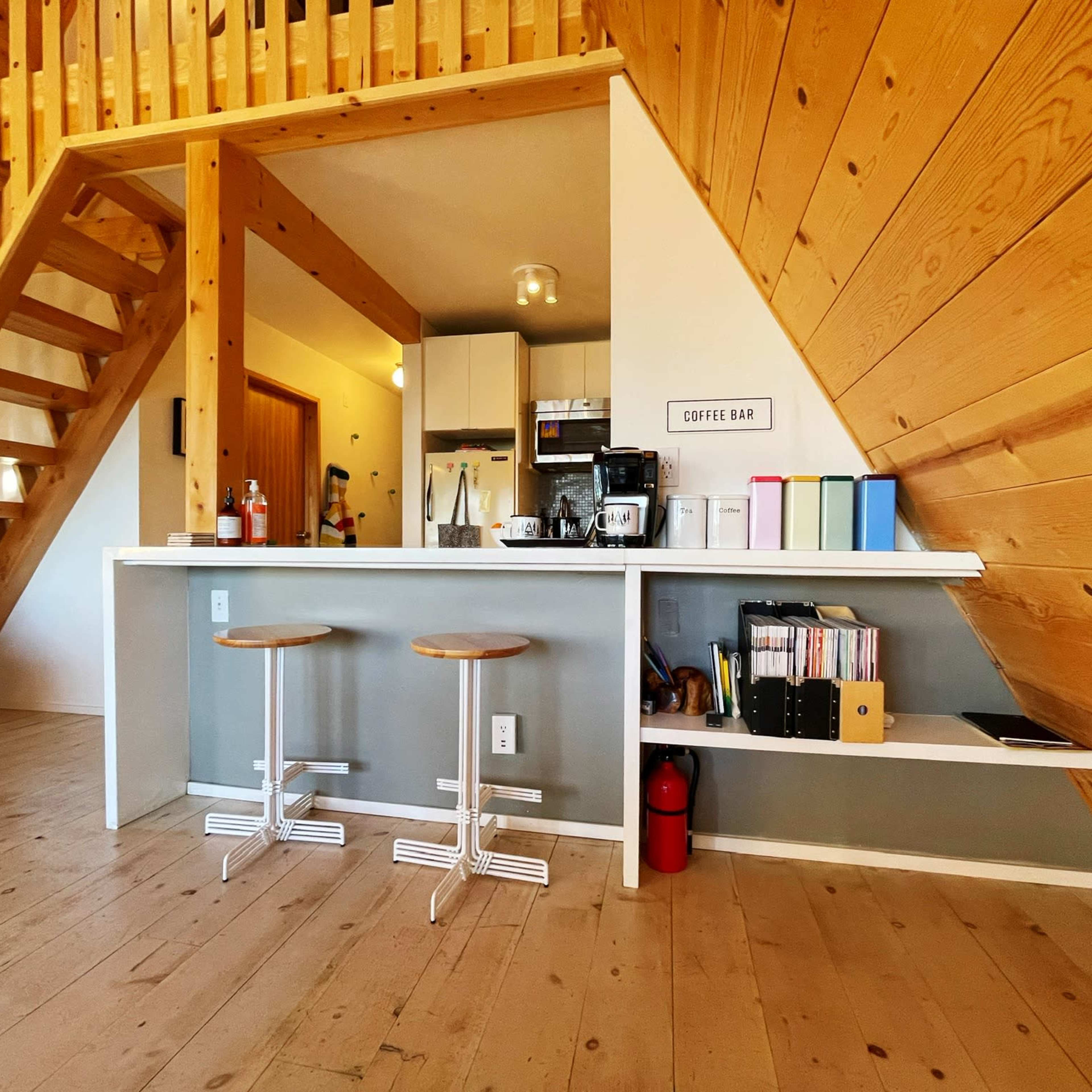 A modern kitchen features a counter with two stools, a coffee bar, and wooden walls alongside a staircase.
