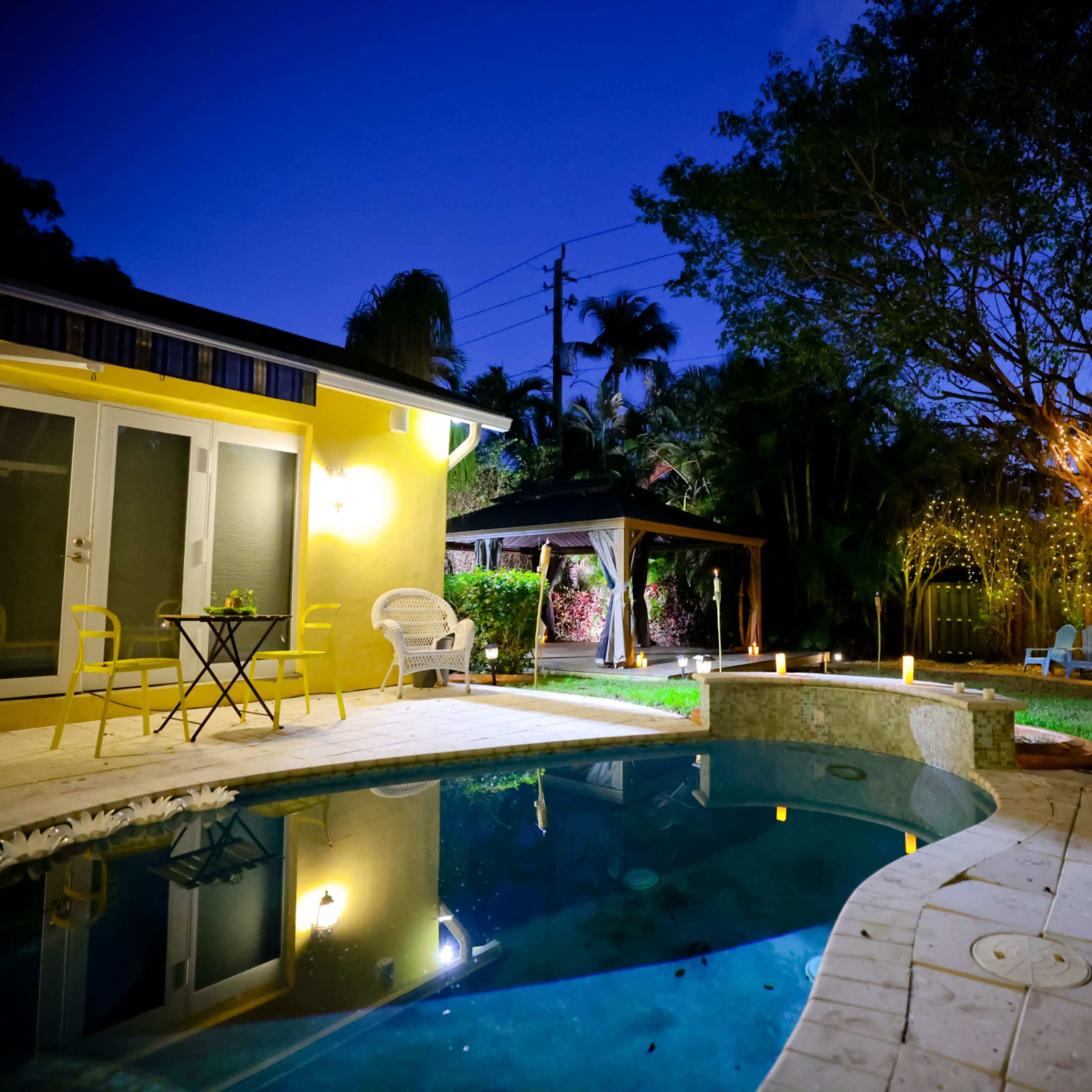 A lit outdoor area featuring a pool, a small table with chairs, and a gazebo surrounded by greenery at dusk.