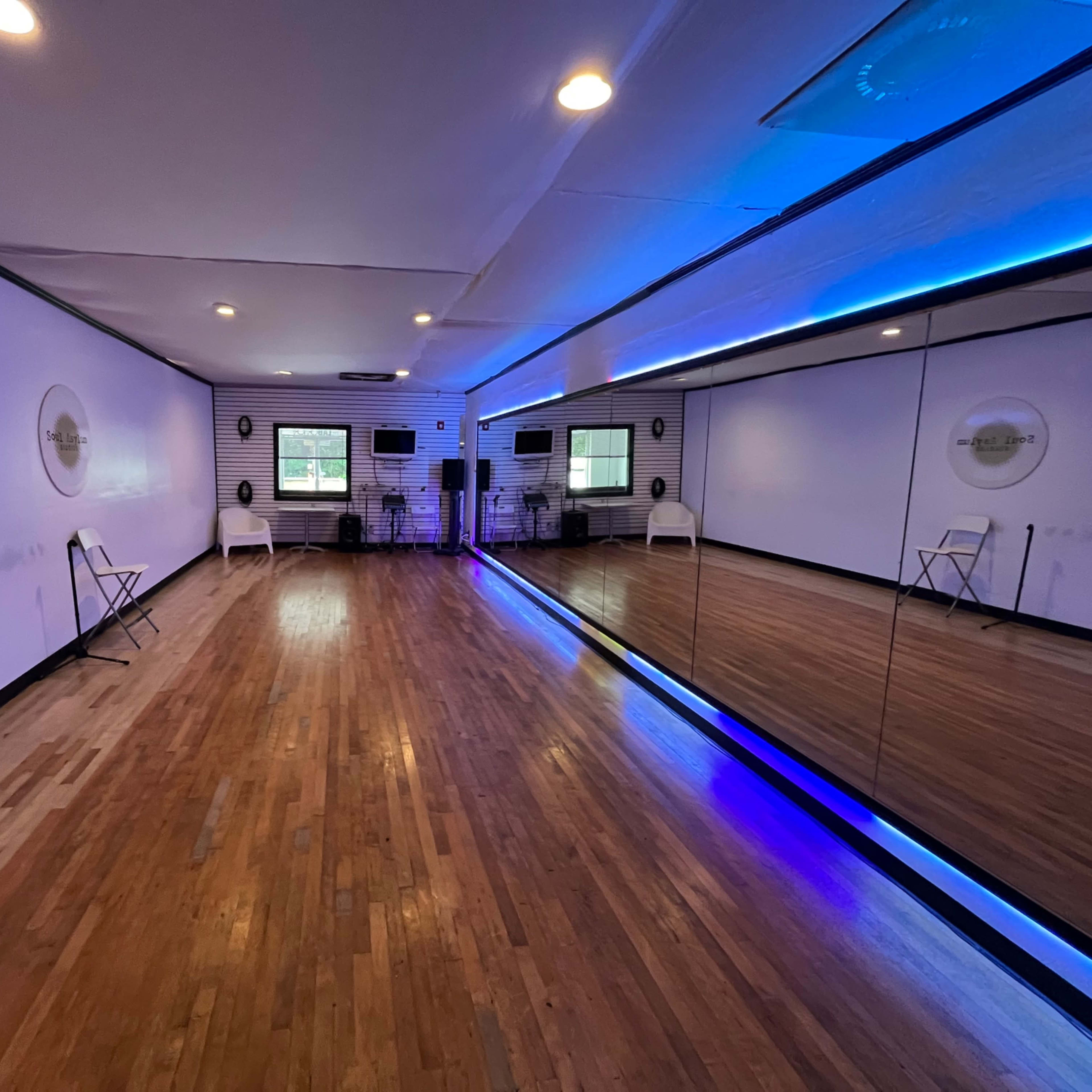 A spacious dance studio features hardwood floors, white walls, and a large mirror with blue lighting along one side.