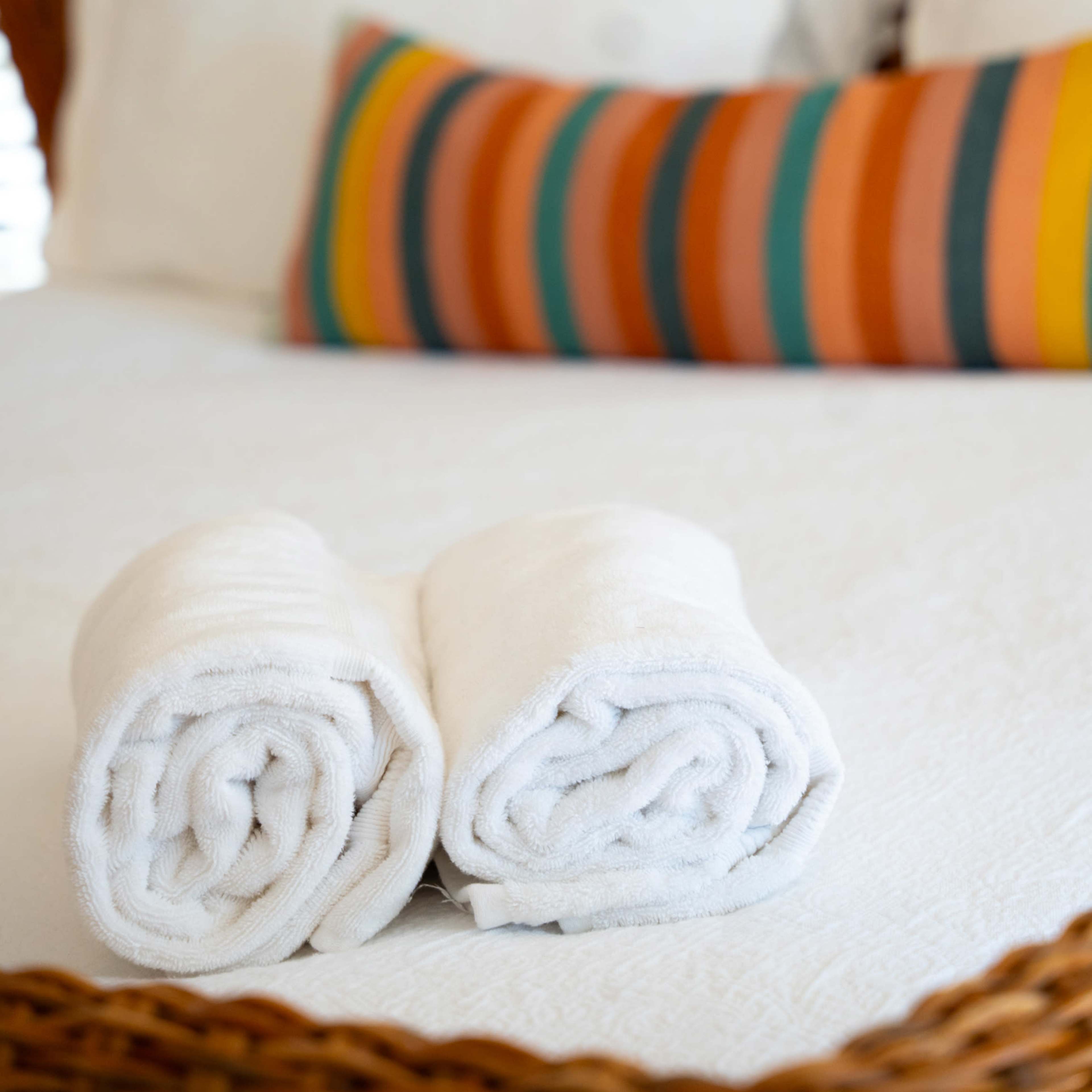 Two rolled white towels are placed on a neatly made bed with a striped decorative pillow in the background.
