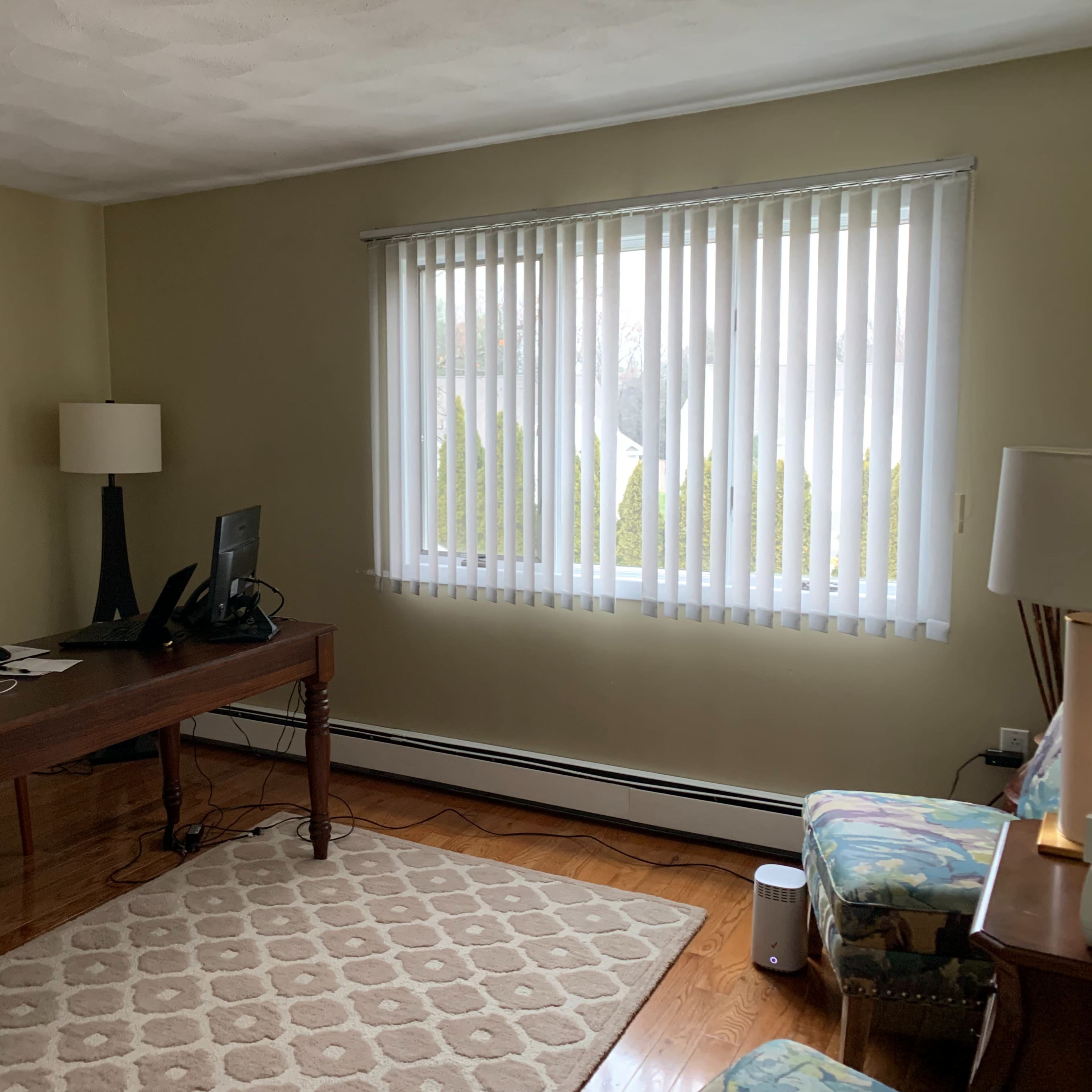 A well-lit office space features a desk with a computer, a chair, a lamp, and a window covered with blinds, alongside a patterned rug and a colorful armchair.