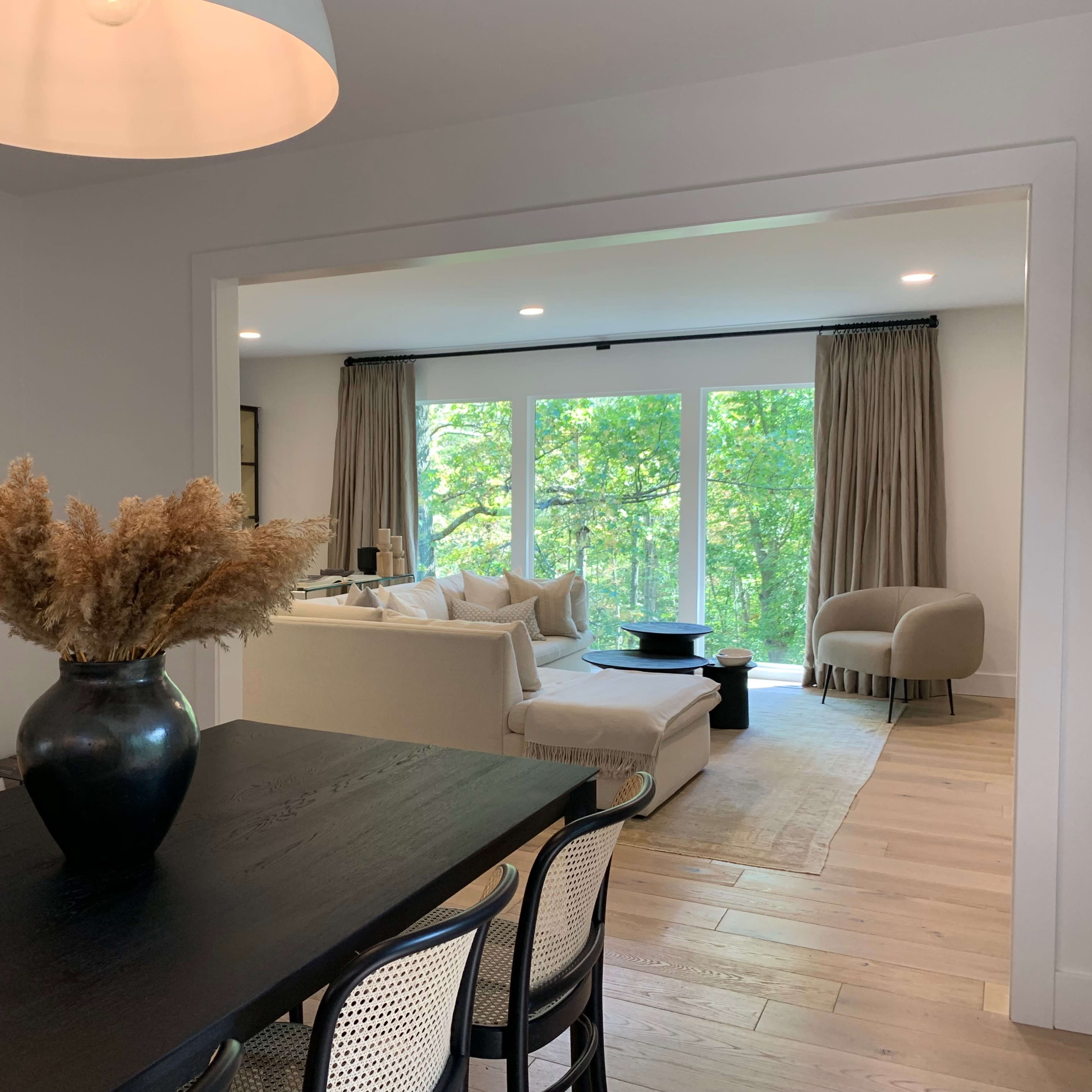 A dining area with a dark wooden table and a vase of dried plants opens into a living room featuring a light-colored sofa and large windows overlooking greenery.