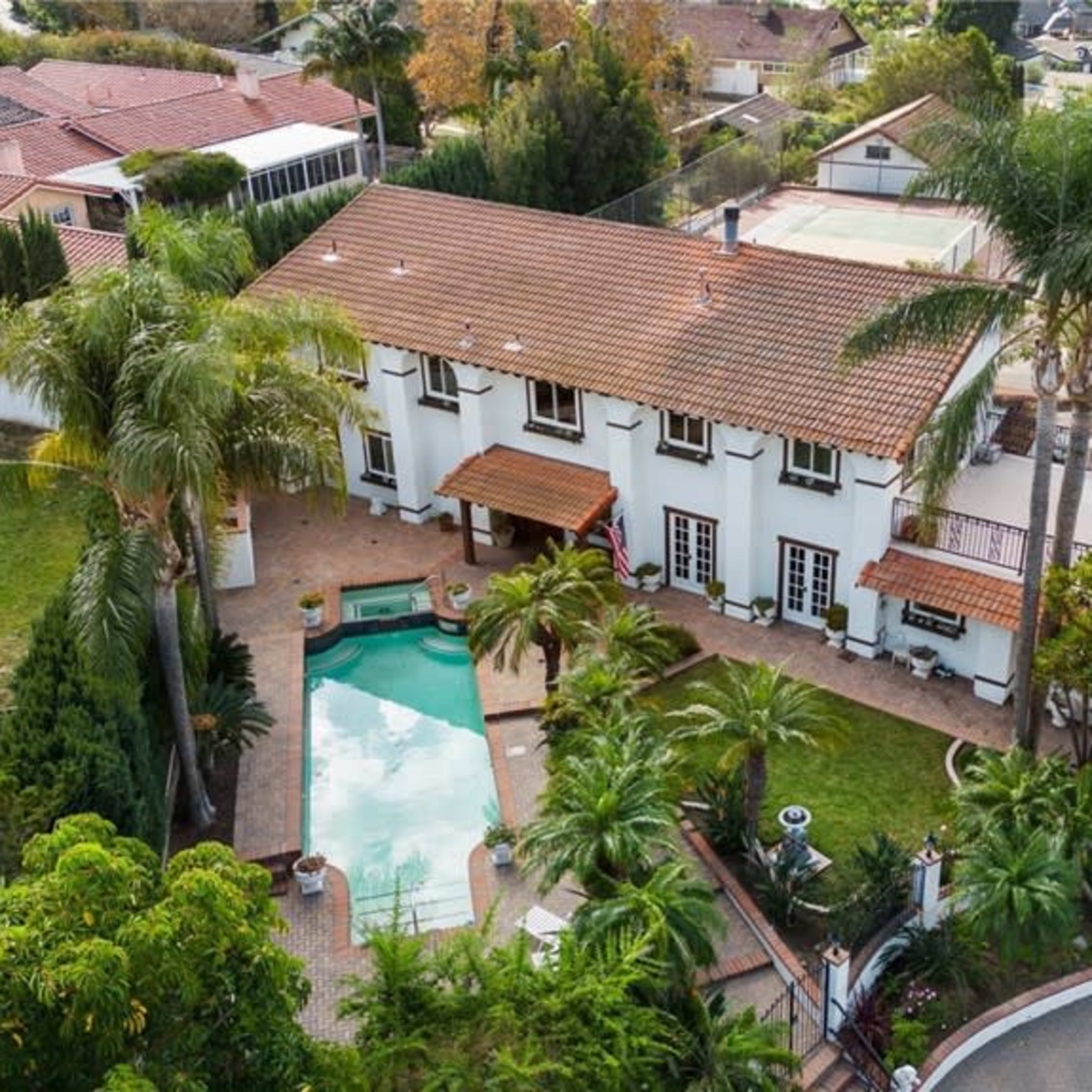 A two-story house with a terracotta tile roof, a swimming pool in the backyard, and surrounding palm trees and landscaped greenery.