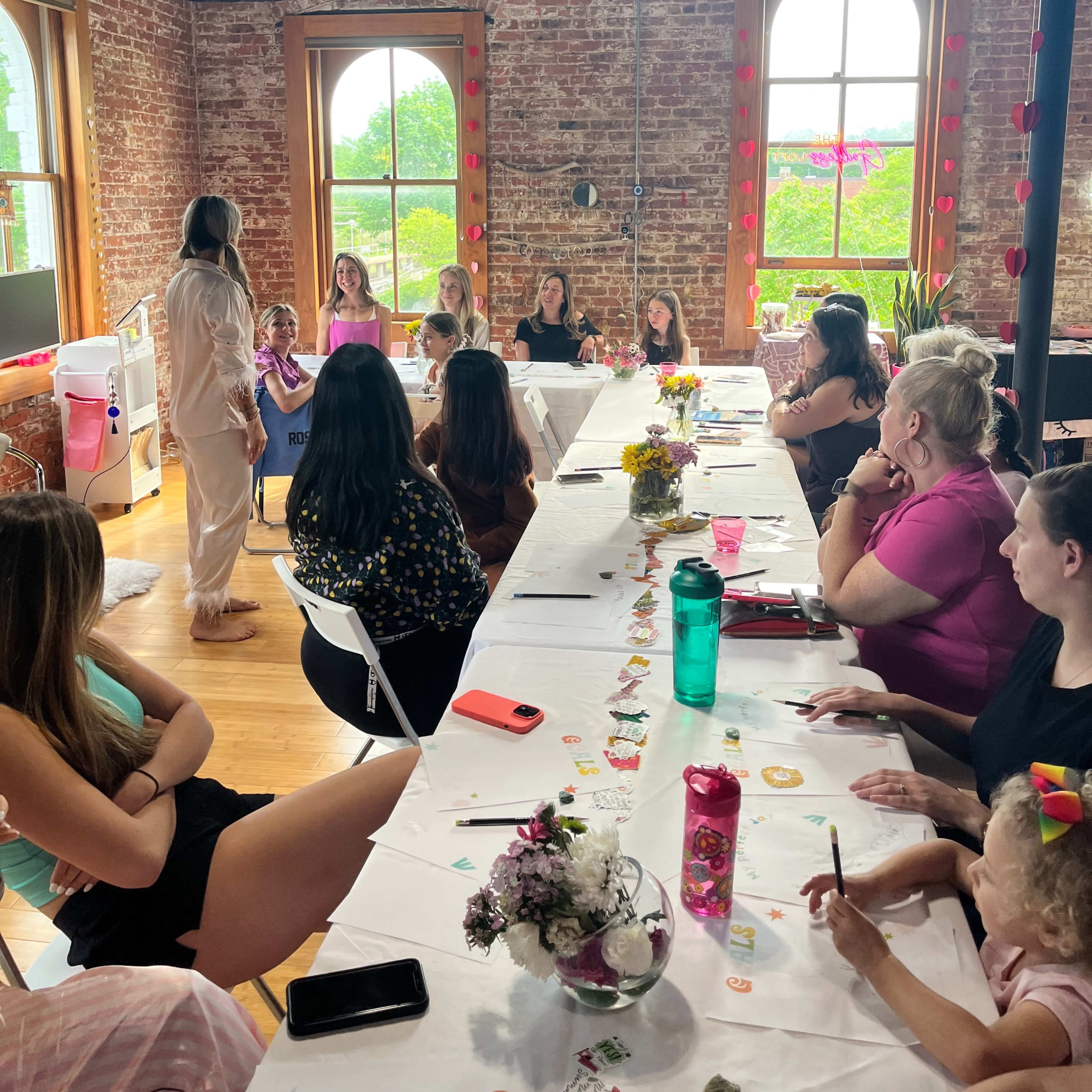 A group of women and children gather around a long table in a room with brick walls and large windows, engaging in a discussion or activity.
