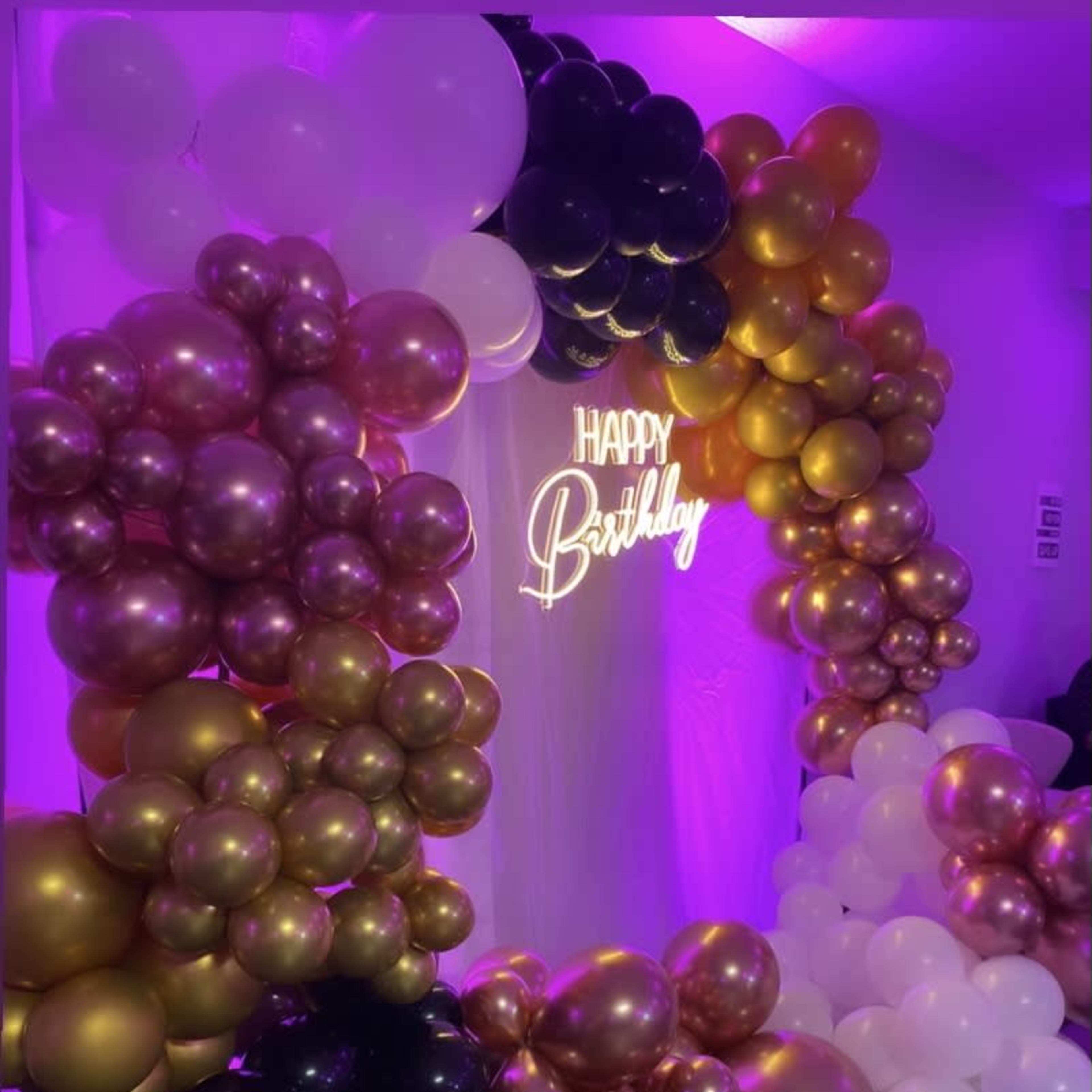 A decorative backdrop for a birthday celebration featuring an arrangement of balloons in gold, black, and white, with a illuminated "Happy Birthday" sign.