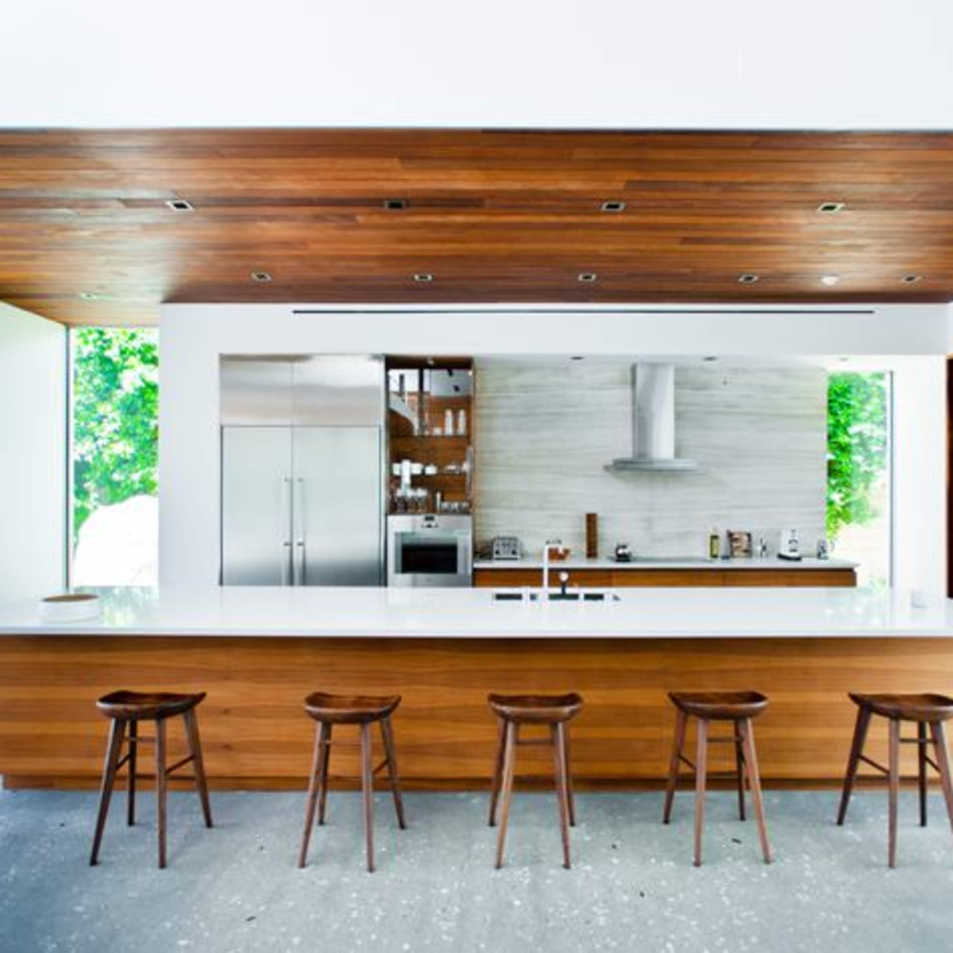 A modern kitchen features a large island with wooden stools, stainless steel appliances, and an open layout highlighted by natural light.