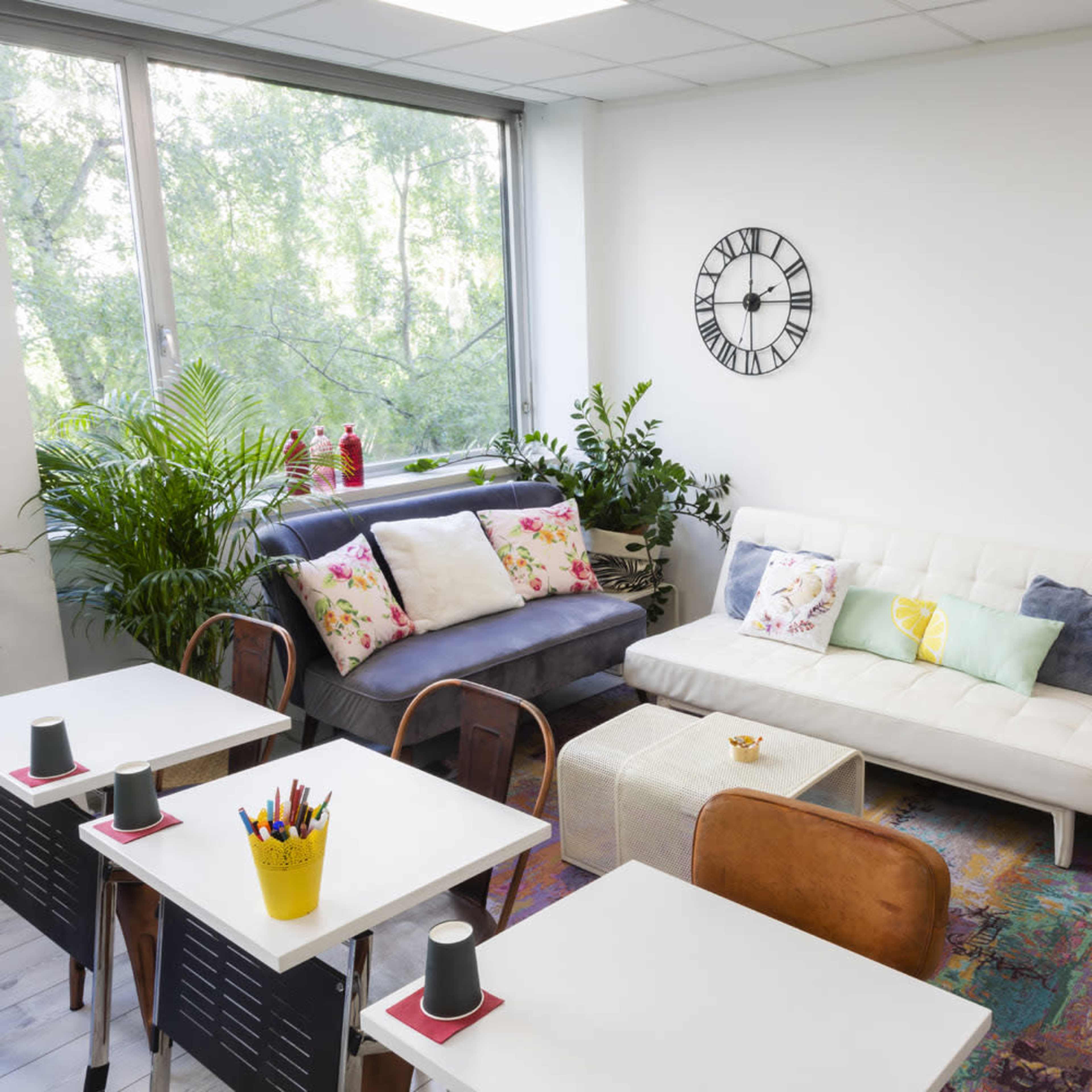 A bright, modern lounge area with two sofas, a coffee table, potted plants, and a wall clock.