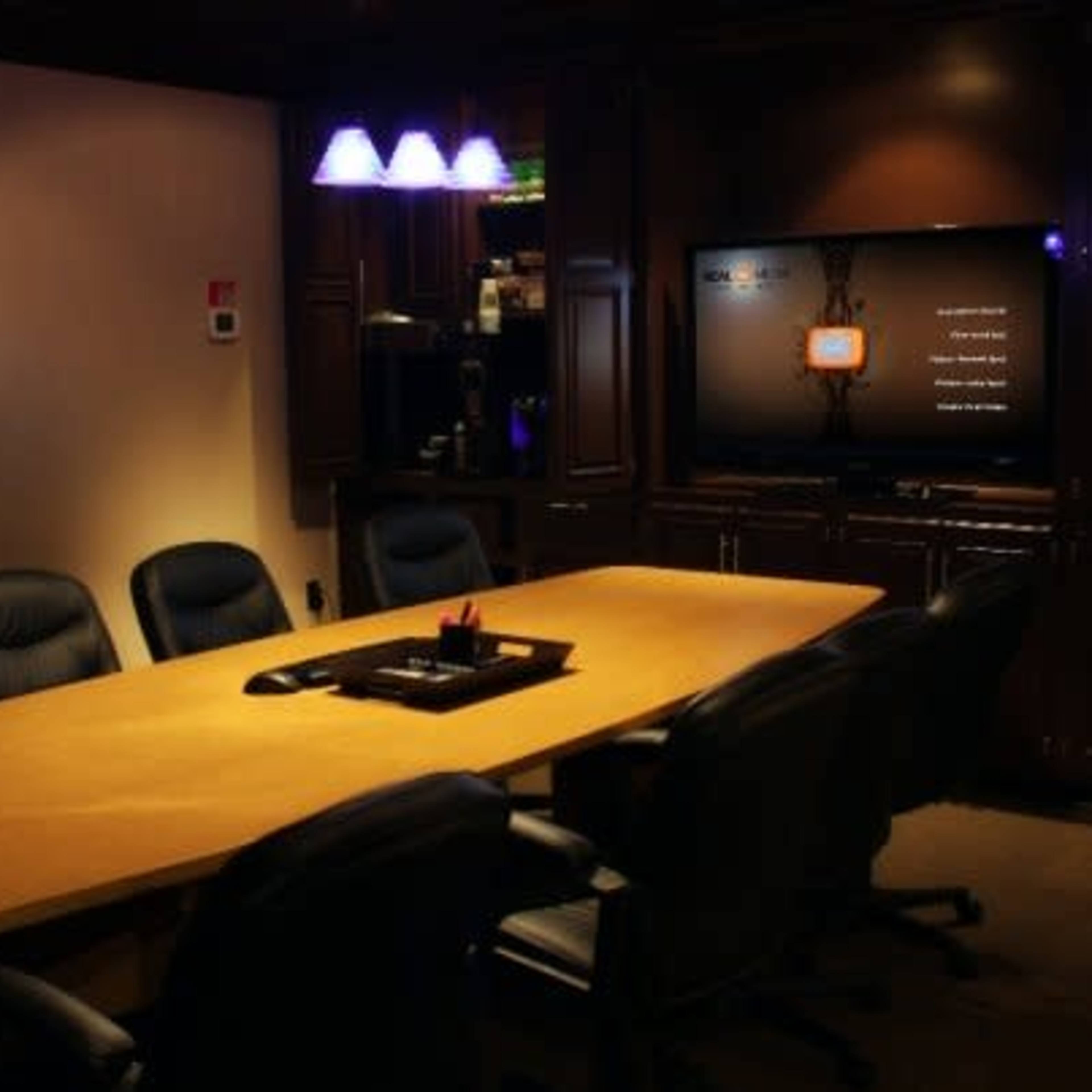 A large wooden conference table surrounded by black chairs sits in a dimly lit boardroom with a television screen displaying a menu on the wall.