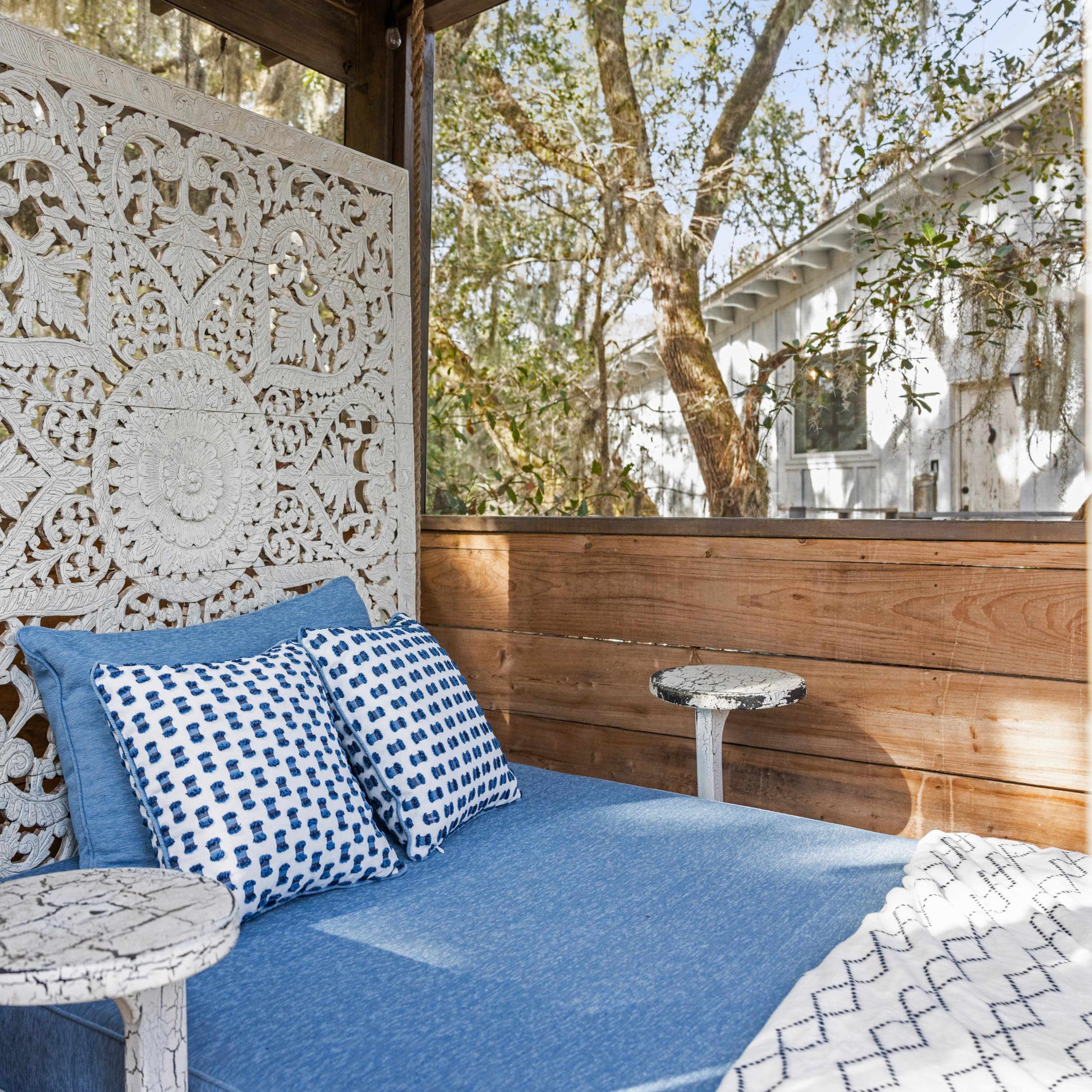 A cozy outdoor seating area featuring a blue upholstered daybed with patterned pillows and intricate white wooden panels in the background.