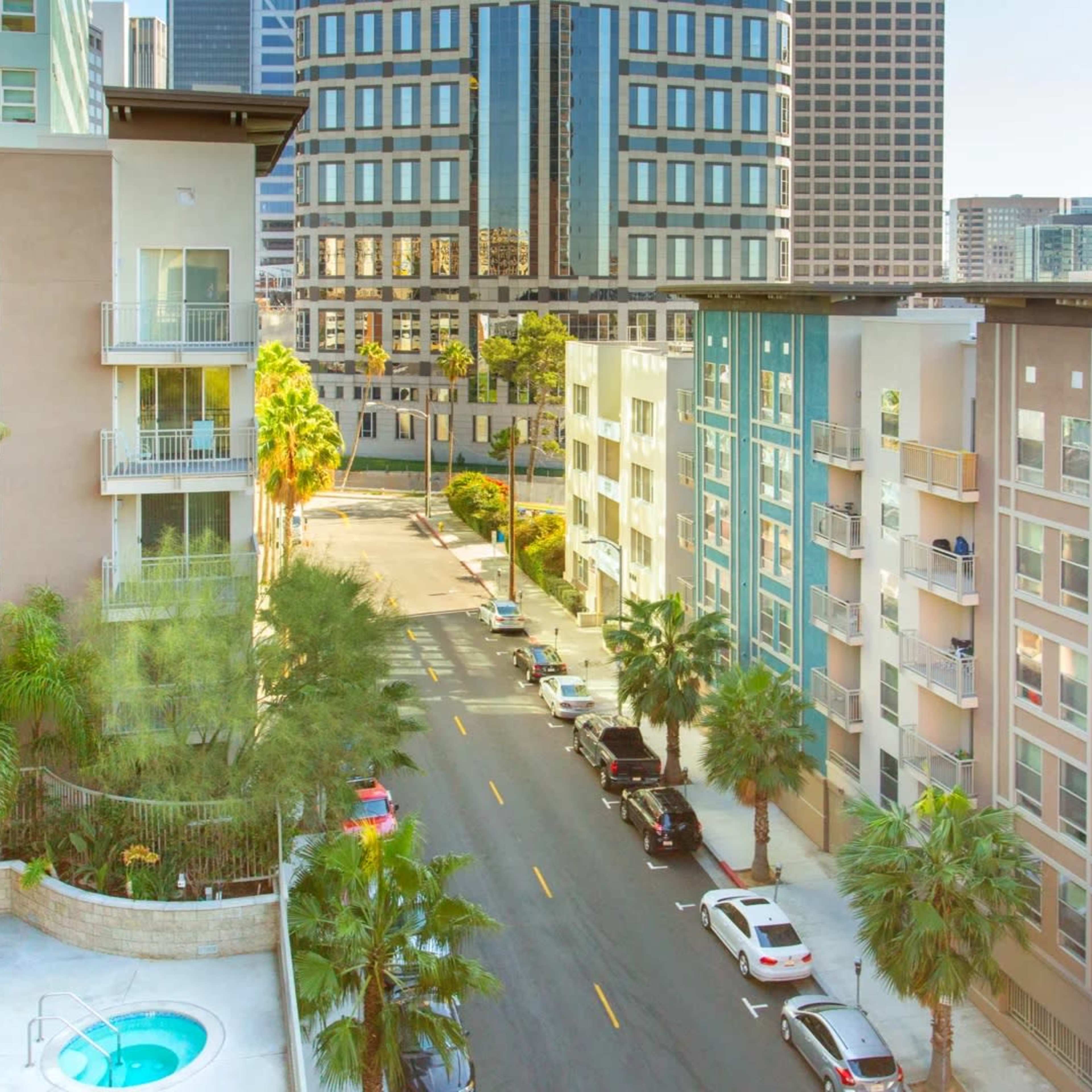 A city street lined with modern apartment buildings and palm trees, with cars parked along the road.
